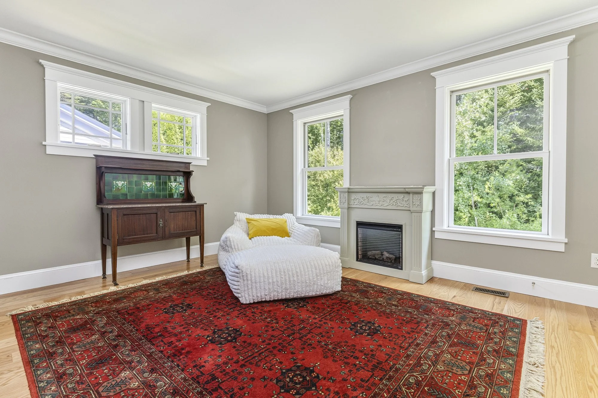 Living room with a red patterned rug, white armchair with a yellow pillow, a dark wooden cabinet with green tiles, white fireplace, and three large windows showing green trees outside.