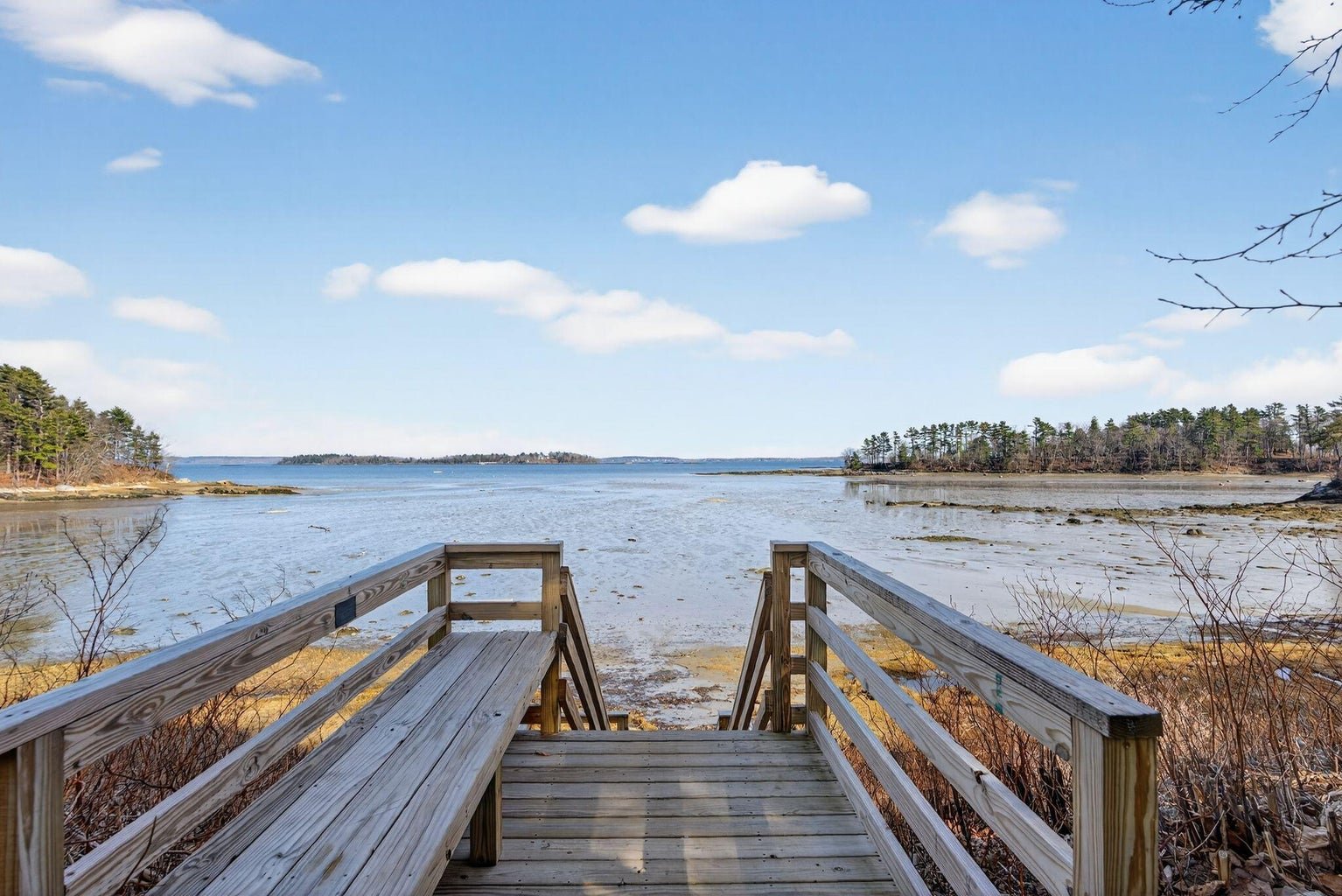 Wooden stairs leading down to a muddy shoreline overlooking a calm body of water with distant trees and a partly cloudy sky.