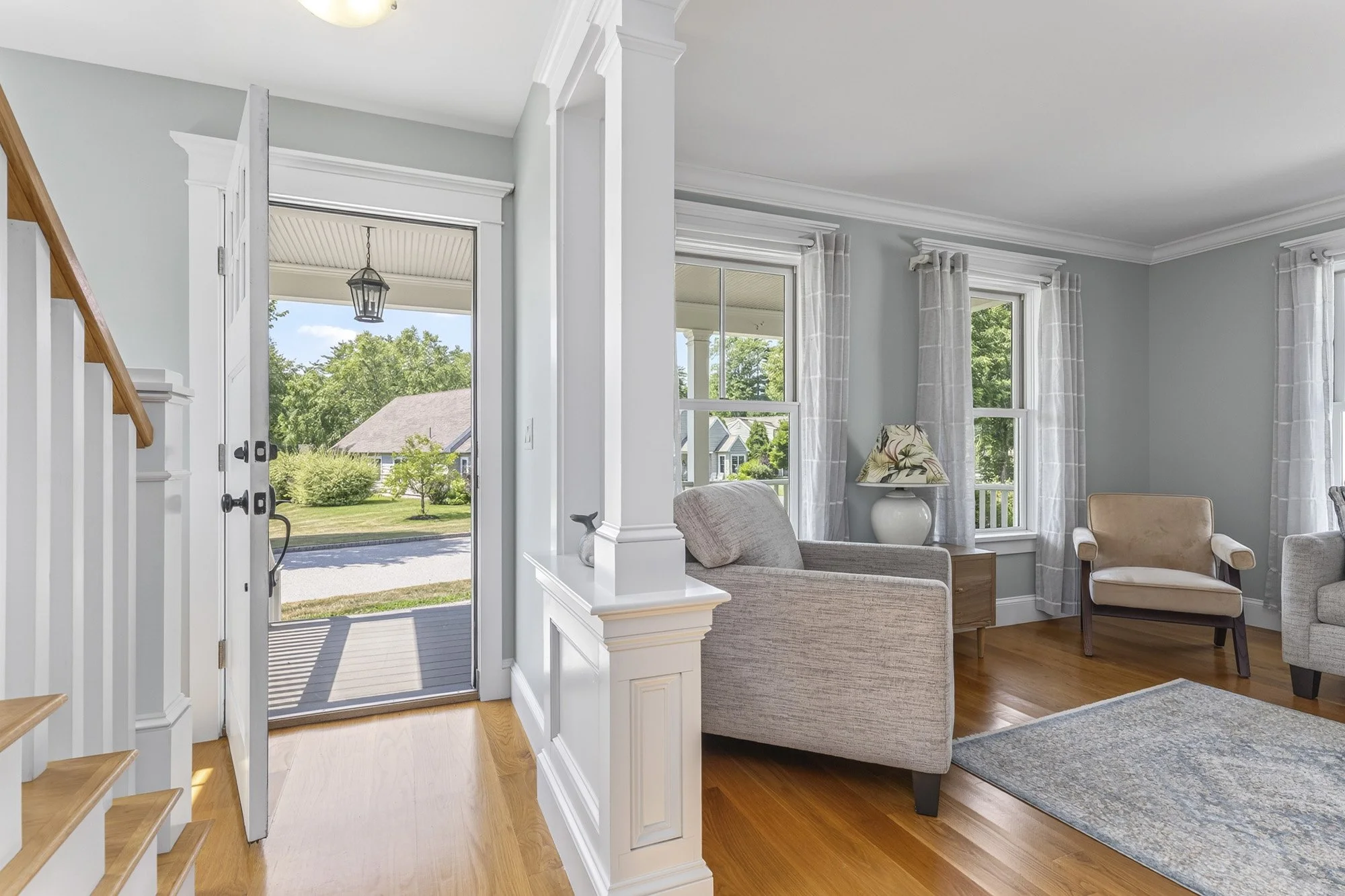 Living room with light gray walls, white trim, hardwood floors, a beige armchair, a gray sofa with pillows, a hanging lamp outside, and large windows with curtains showing a green outdoor yard.
