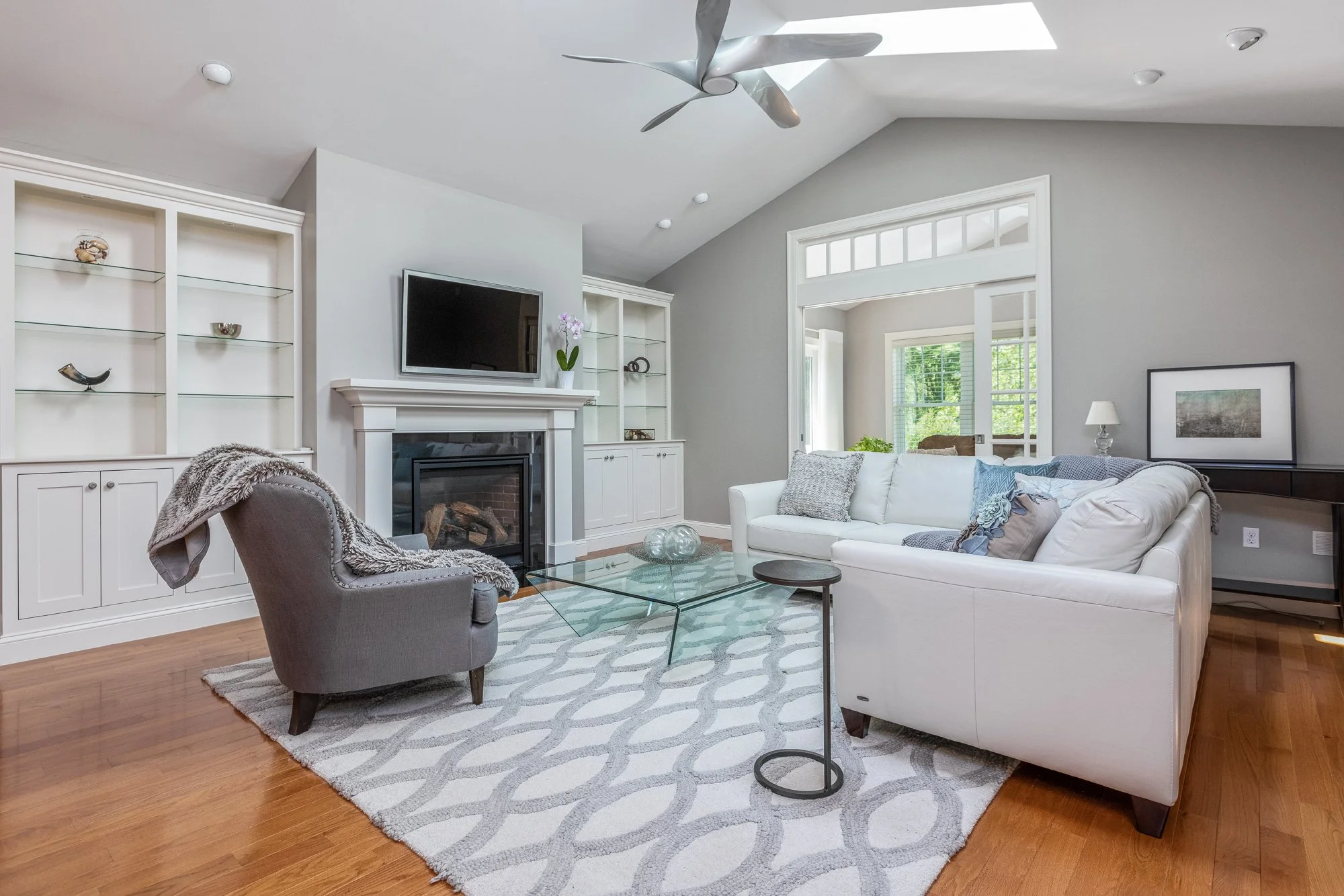 Living room with a white sofa, gray armchair with a fuzzy throw, glass coffee table, area rug, built-in bookshelves, fireplace, flat-screen TV, potted orchid, and framed artwork, with windows and skylight allowing natural light.