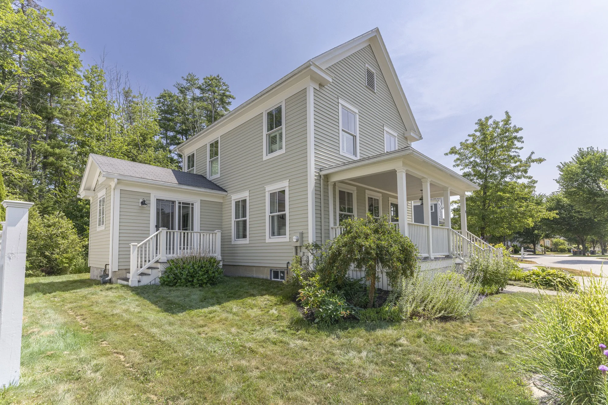 A two-story light yellow house with white trim, a porch with white railings, surrounded by a well-kept lawn and garden, with trees in the background and a clear blue sky.