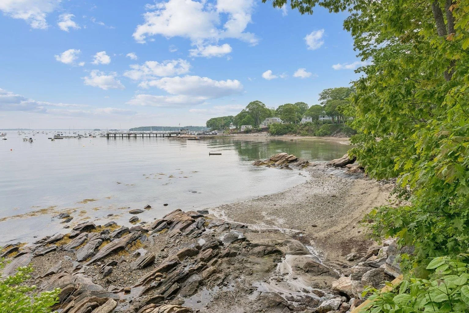 Calm coastal scene with rocky shoreline, sandy beach, green trees, houses on a hill, and a pier with boats on a partly cloudy day.