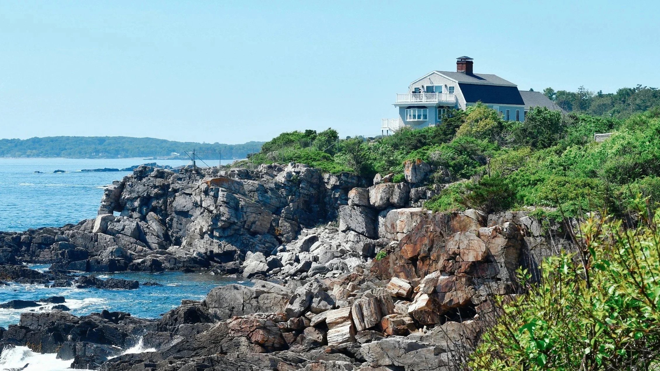 A house perched on rocky cliffs overlooking a body of water with trees in the distance.