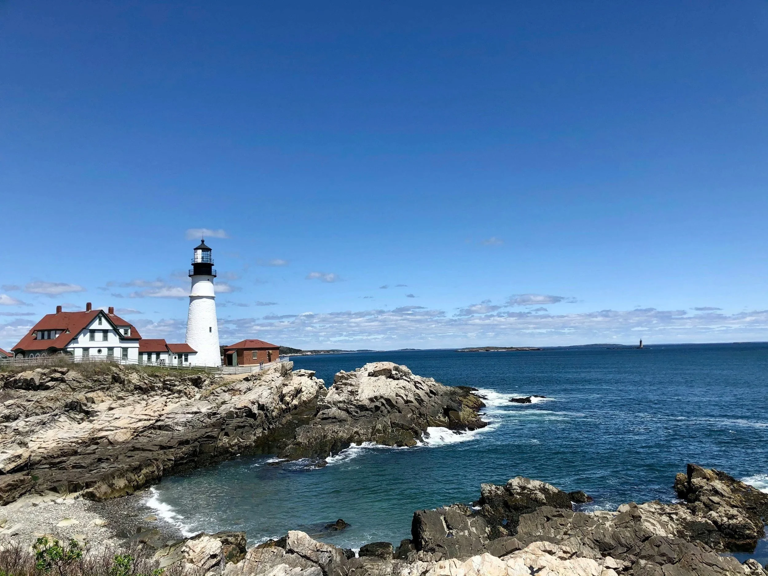 A lighthouse near the rocky coastline, with a house nearby, under a bright blue sky with some scattered clouds.