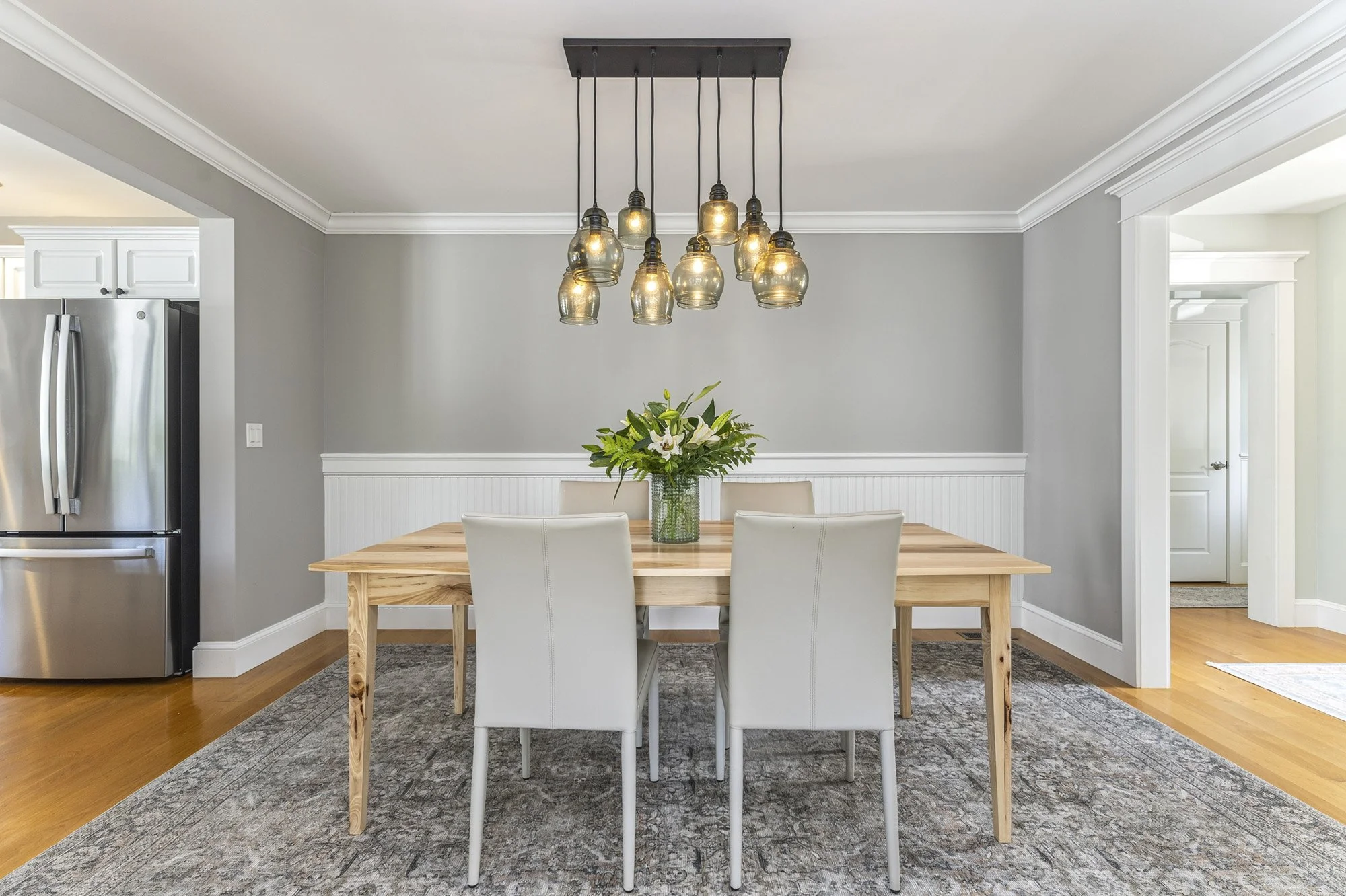 A contemporary dining room featuring a wooden table with light-colored chairs, a floral centerpiece, a modern multi-bulb pendant light fixture, and a kitchen area with a stainless steel refrigerator in the background.