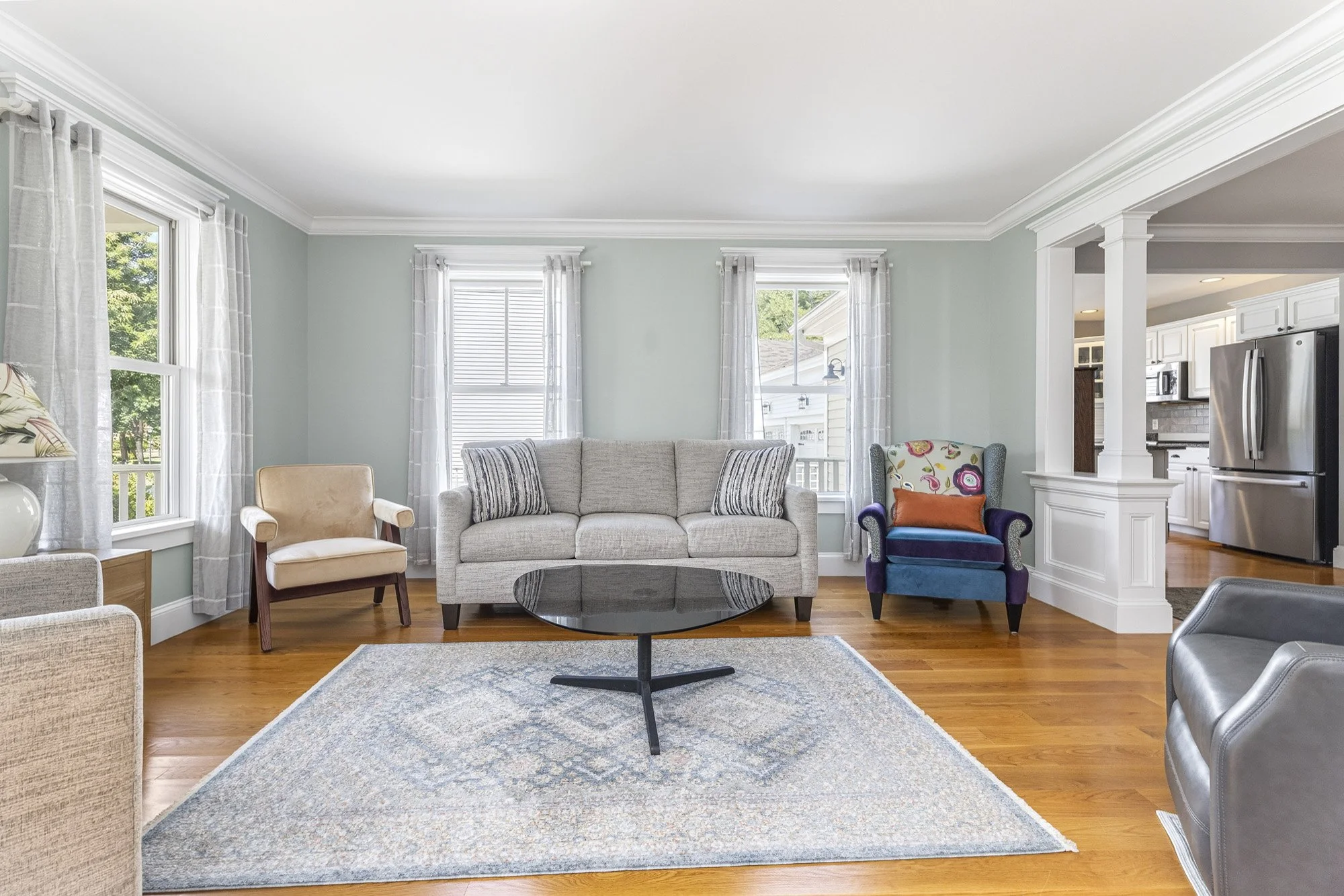 Living room with light green walls, large windows with white curtains, a beige armchair, a beige sofa with striped pillows, a blue and multicolor armchair, a black coffee table, and wooden flooring. Part of kitchen area with white cabinets and a stainless steel refrigerator is visible.