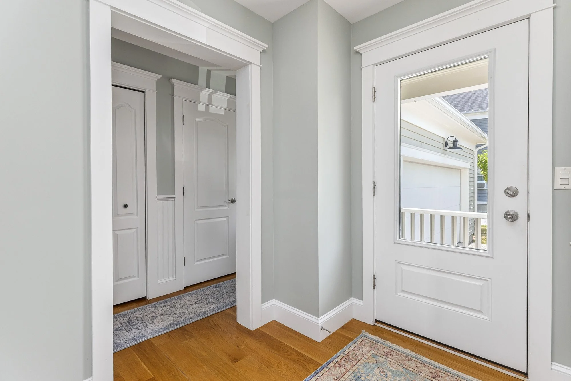 Entryway with a white door leading outside, a window beside the door showing a porch and house exterior, and a gray area rug on hardwood floor.