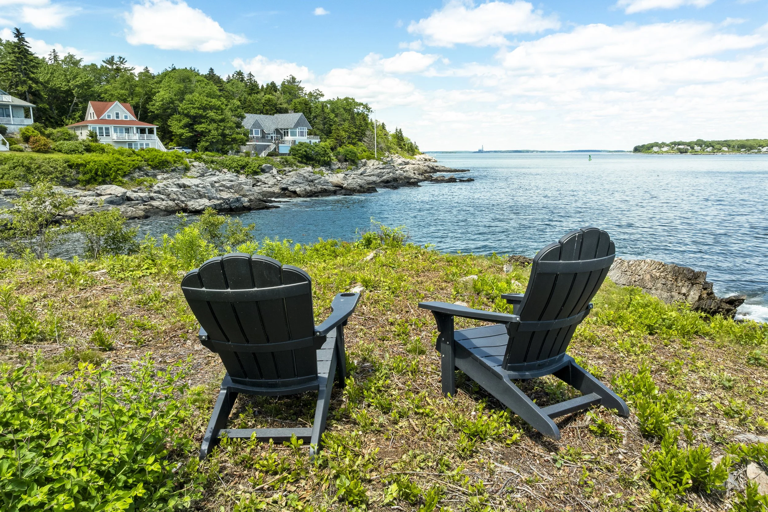 Two black Adirondack chairs placed on grass by a rocky shoreline overlooking Casco Bay Maine from 109 Reed Avenue on Peaks Island with houses and trees in the background under a partly cloudy sky.