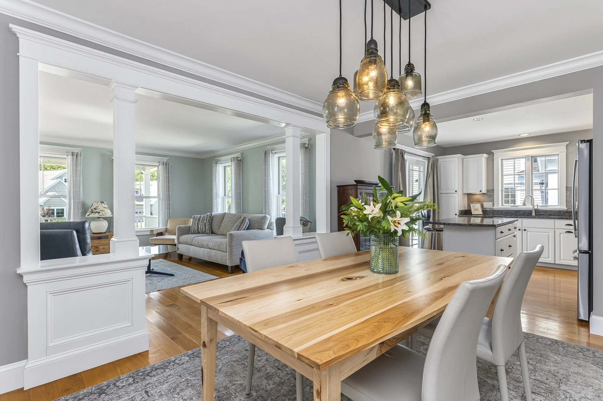 Open-concept living room and kitchen with white cabinetry, light walls, wooden flooring, a wooden dining table with white chairs, a glass vase with lilies, pendant lights, and large windows.