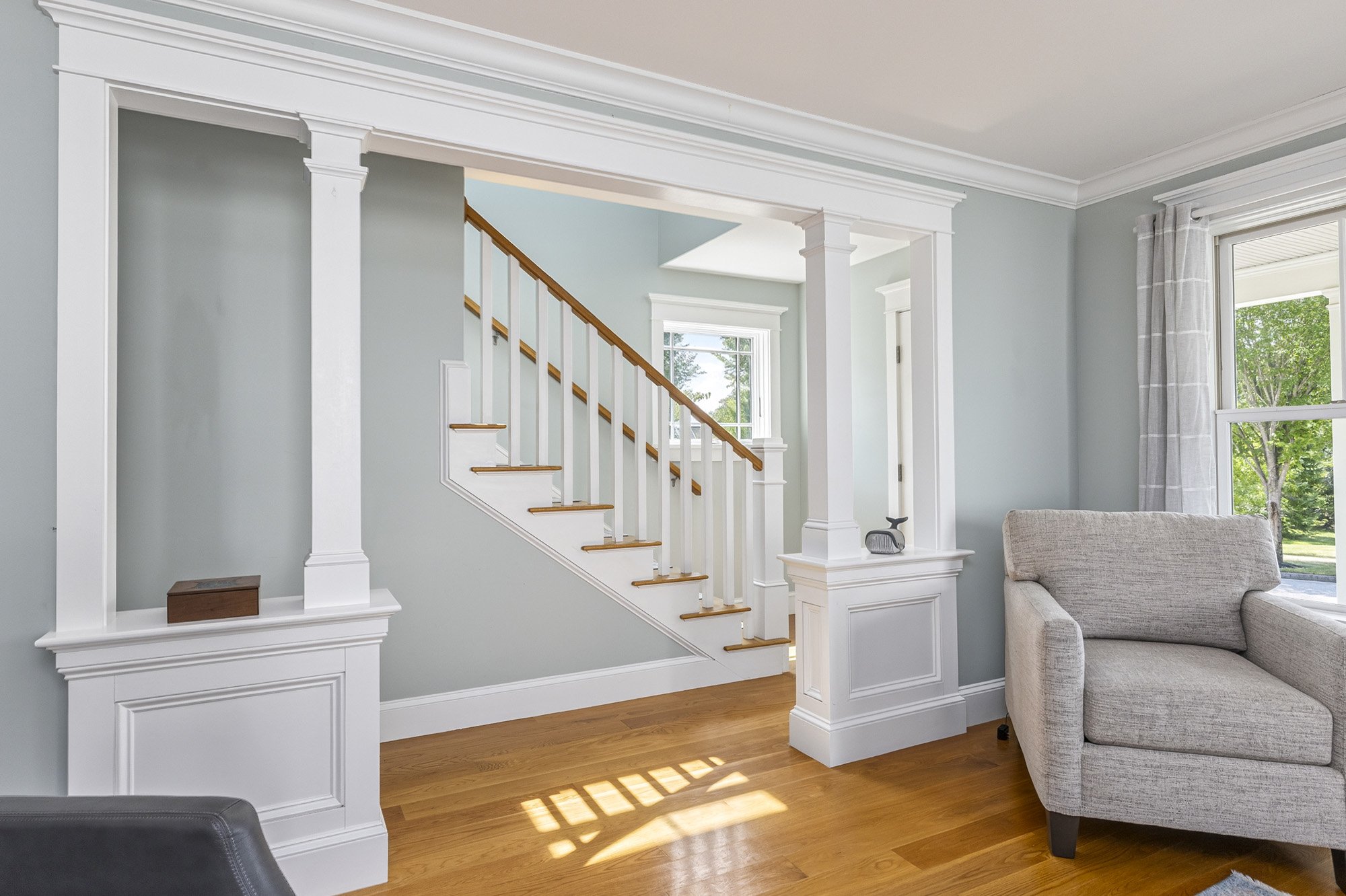 Living room with a staircase, beige sofa, and large windows letting in natural light. Light blue walls and wooden floors.
