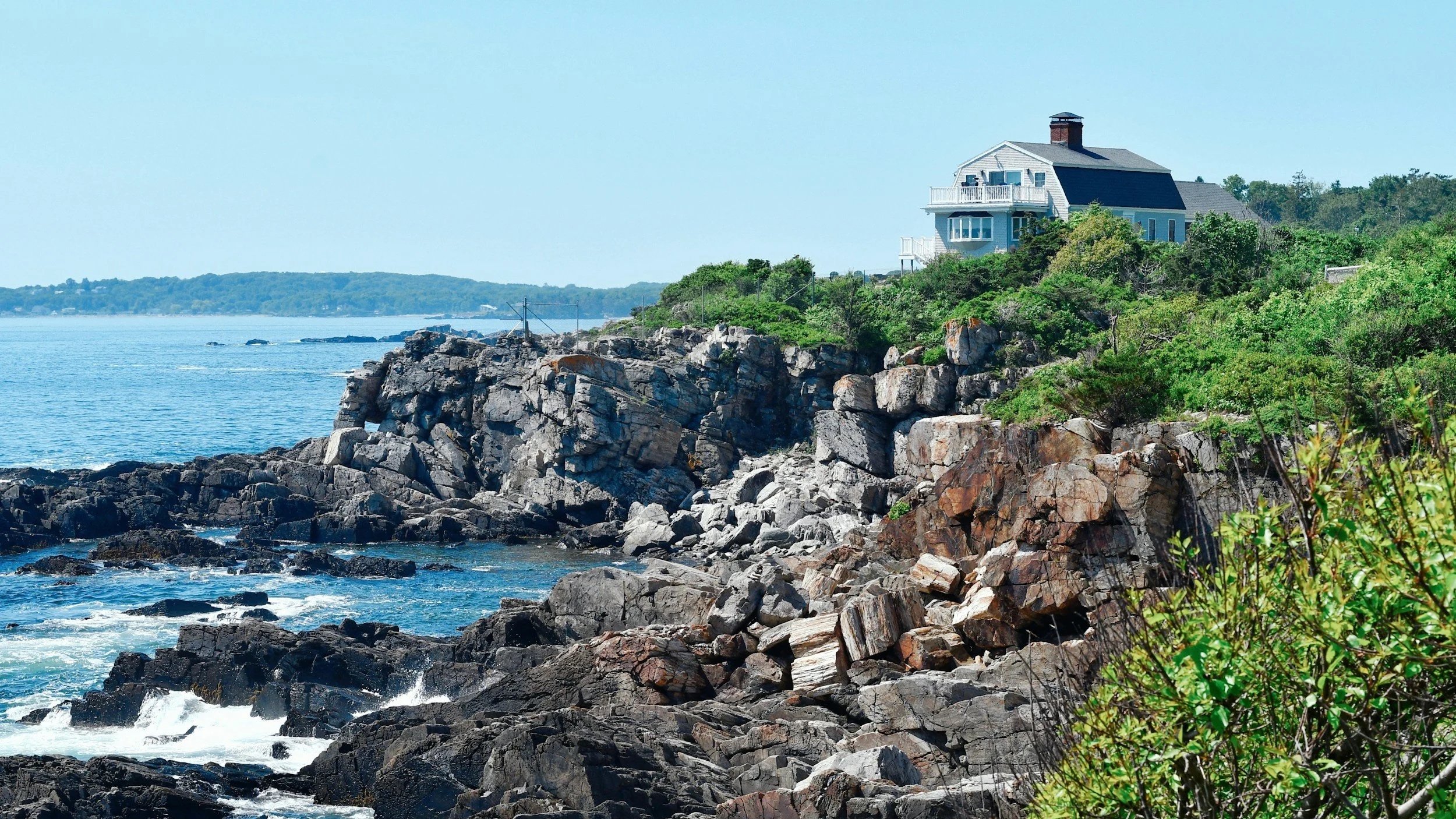A house perched on a rocky coastline overlooking the ocean with green vegetation around.
