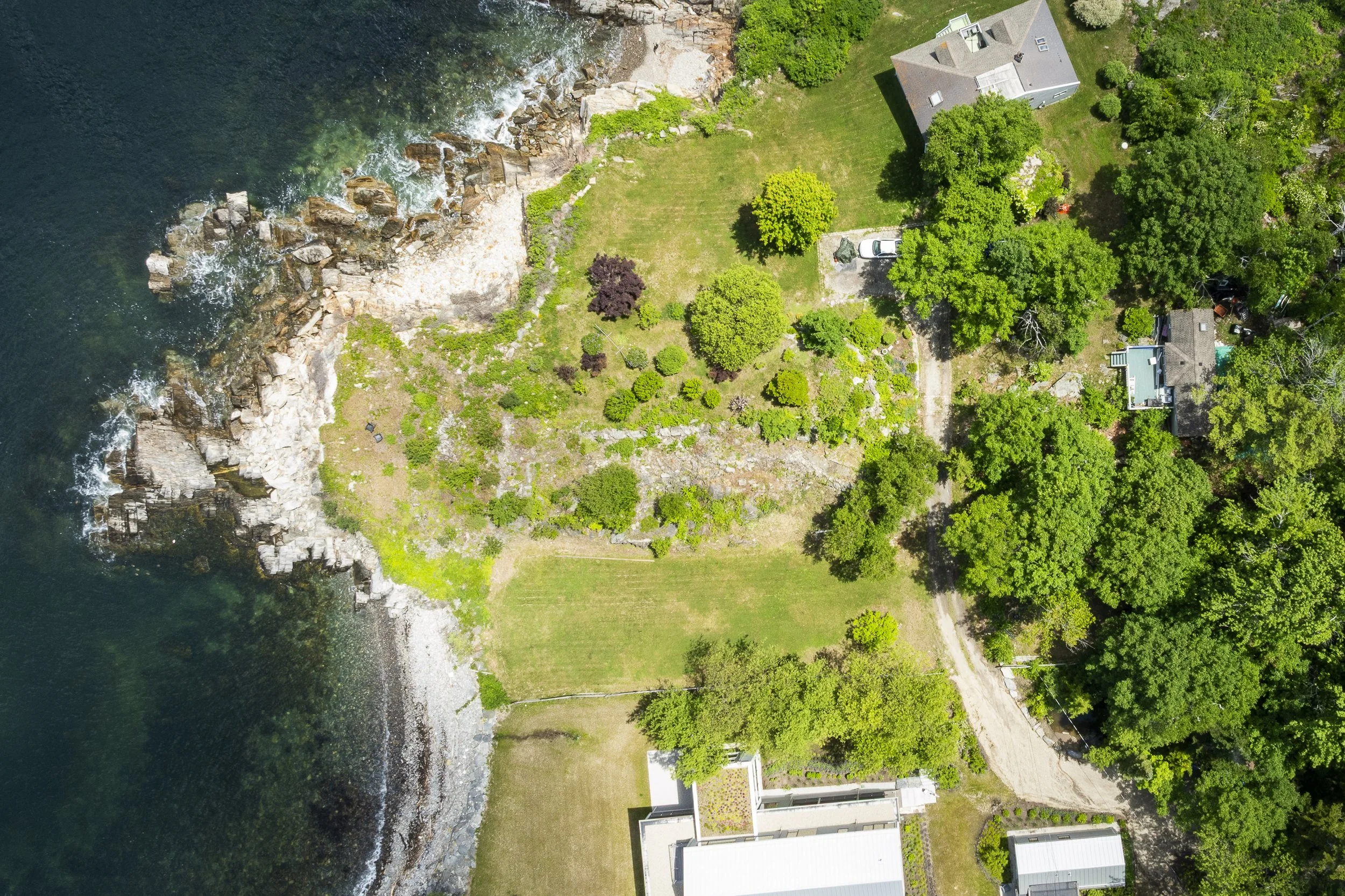 An aerial view of 109 Reed Avenue on Peaks Island, Portland.  Coastal residential area with a large grassy yard, surrounded by trees, adjacent to a rocky shoreline and ocean.