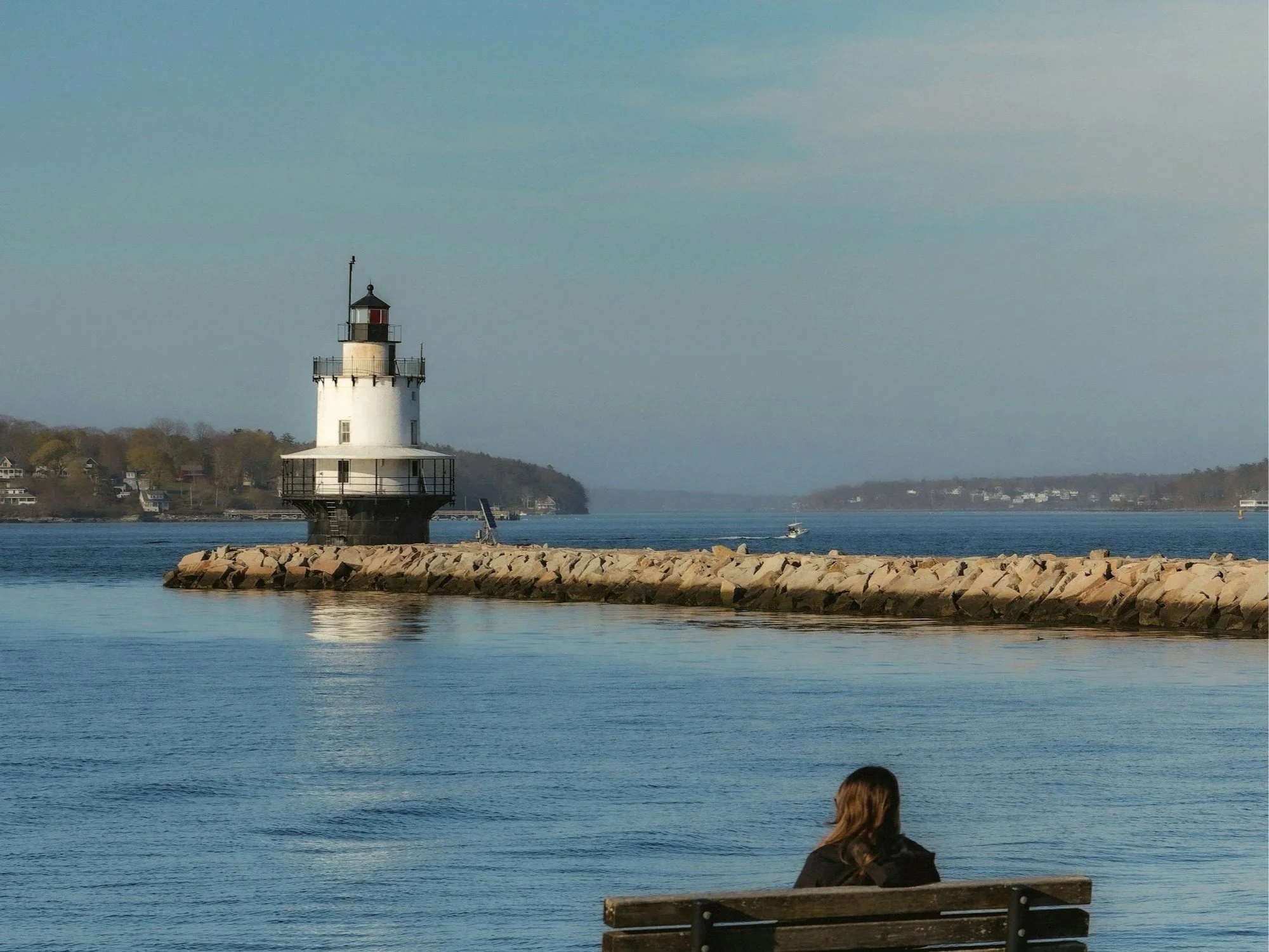 A person sitting on a bench by the water with a lighthouse on a rock jetty in the background, and boats on the water with a distant shoreline on a clear day.