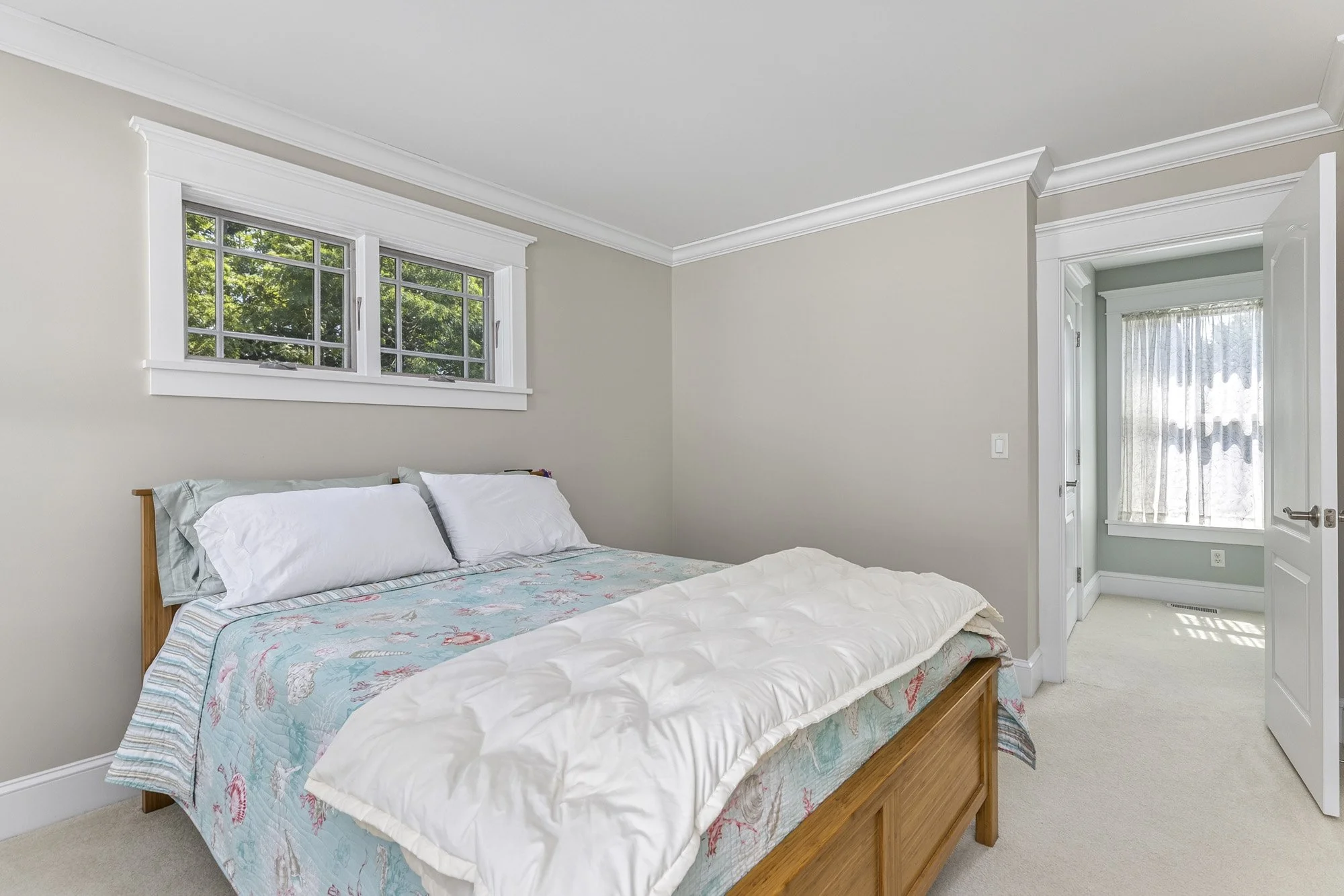 A bedroom with a wooden bed frame, a bed covered with a pastel-colored bedspread featuring marine life, and two white pillows. There are windows with white trim and sheer curtains, and a doorway with a lace curtain at the back.