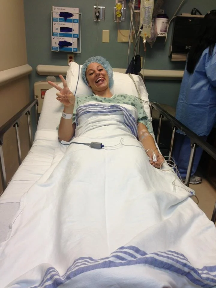 A woman lying in a hospital bed, smiling, wearing a hospital gown and a blue hair cap, making a peace sign with her hand.