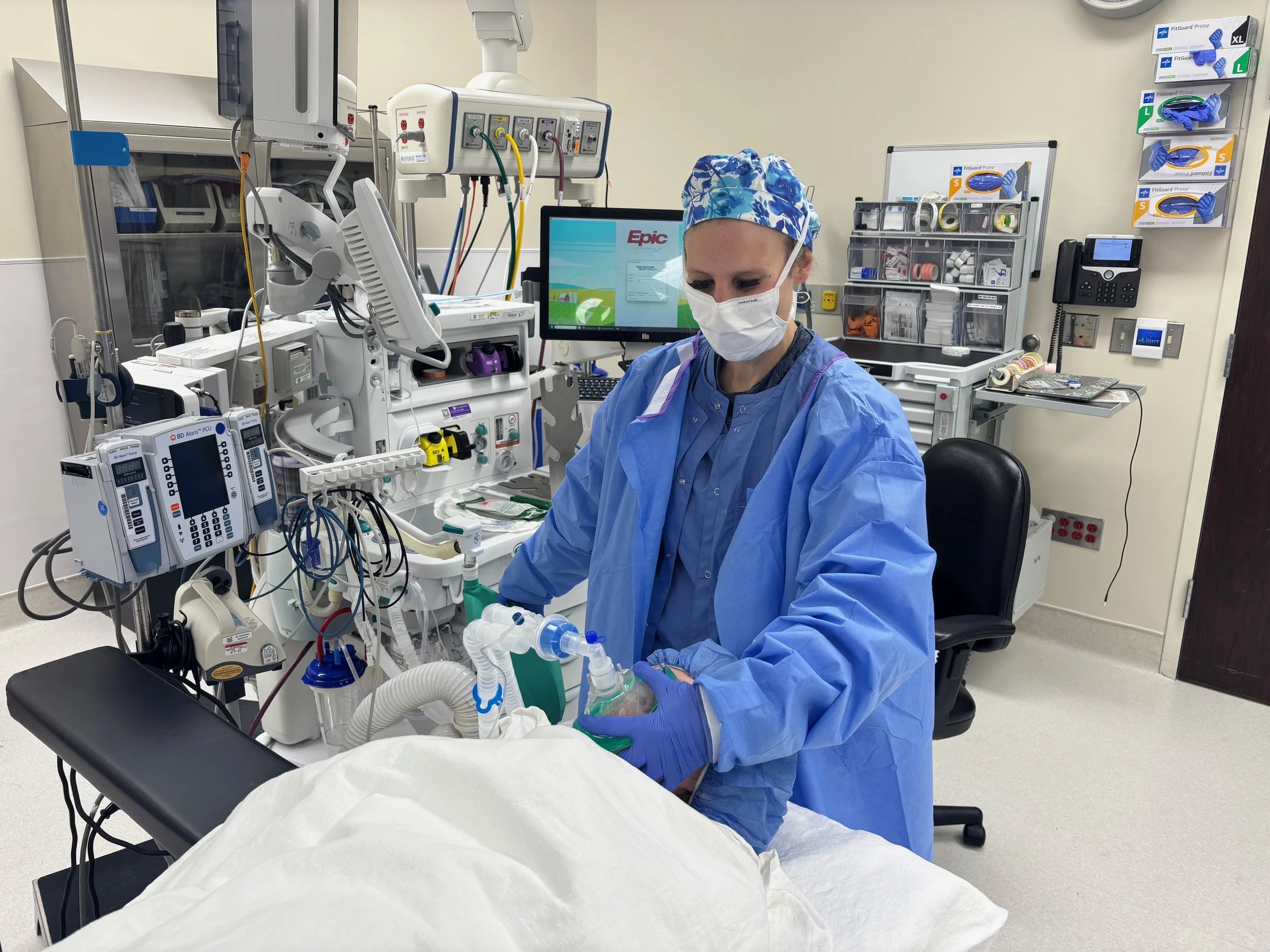 Healthcare professional wearing scrubs, mask, and gloves preparing medical equipment in an intensive care unit.