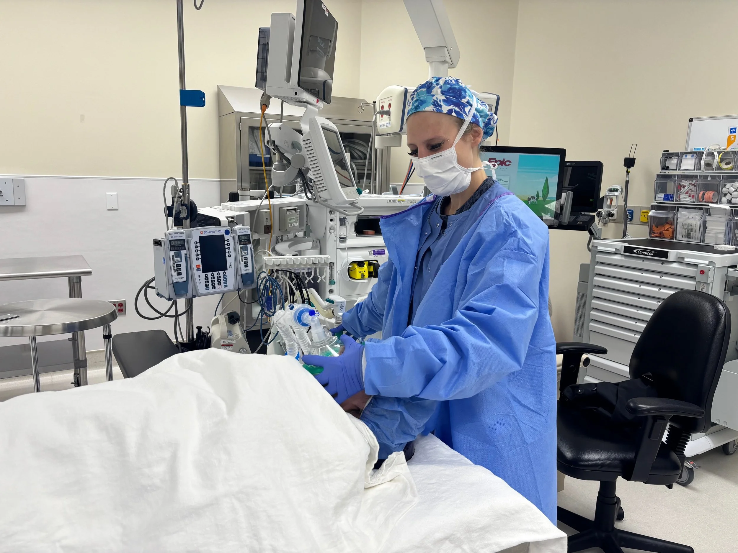 Medical professional in blue scrubs, mask, and gloves adjusts equipment in a hospital room with various medical devices and monitors.