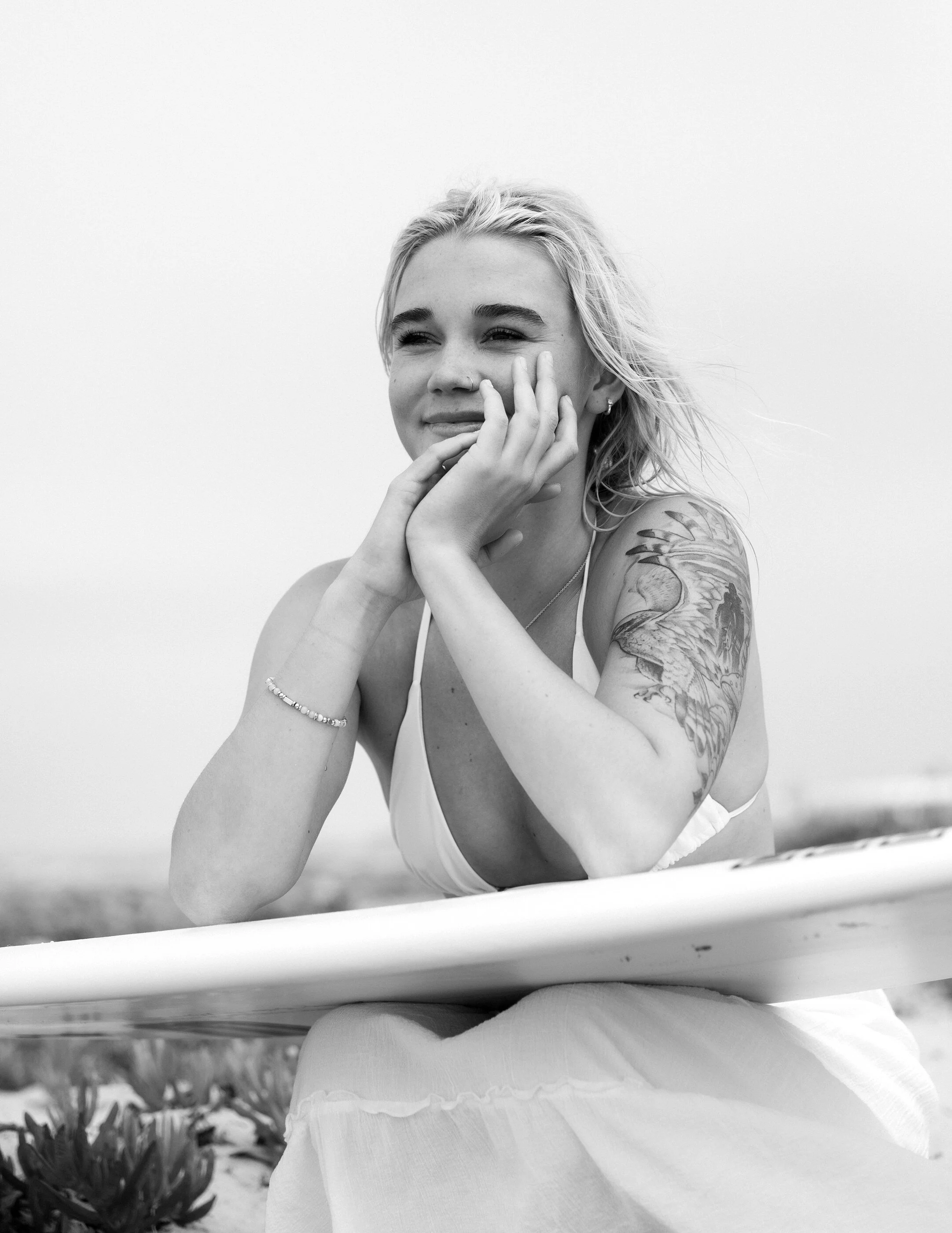 A woman with blonde hair and tattoos lying on her stomach on the beach, resting her chin on her hand, with a surfboard in front of her.