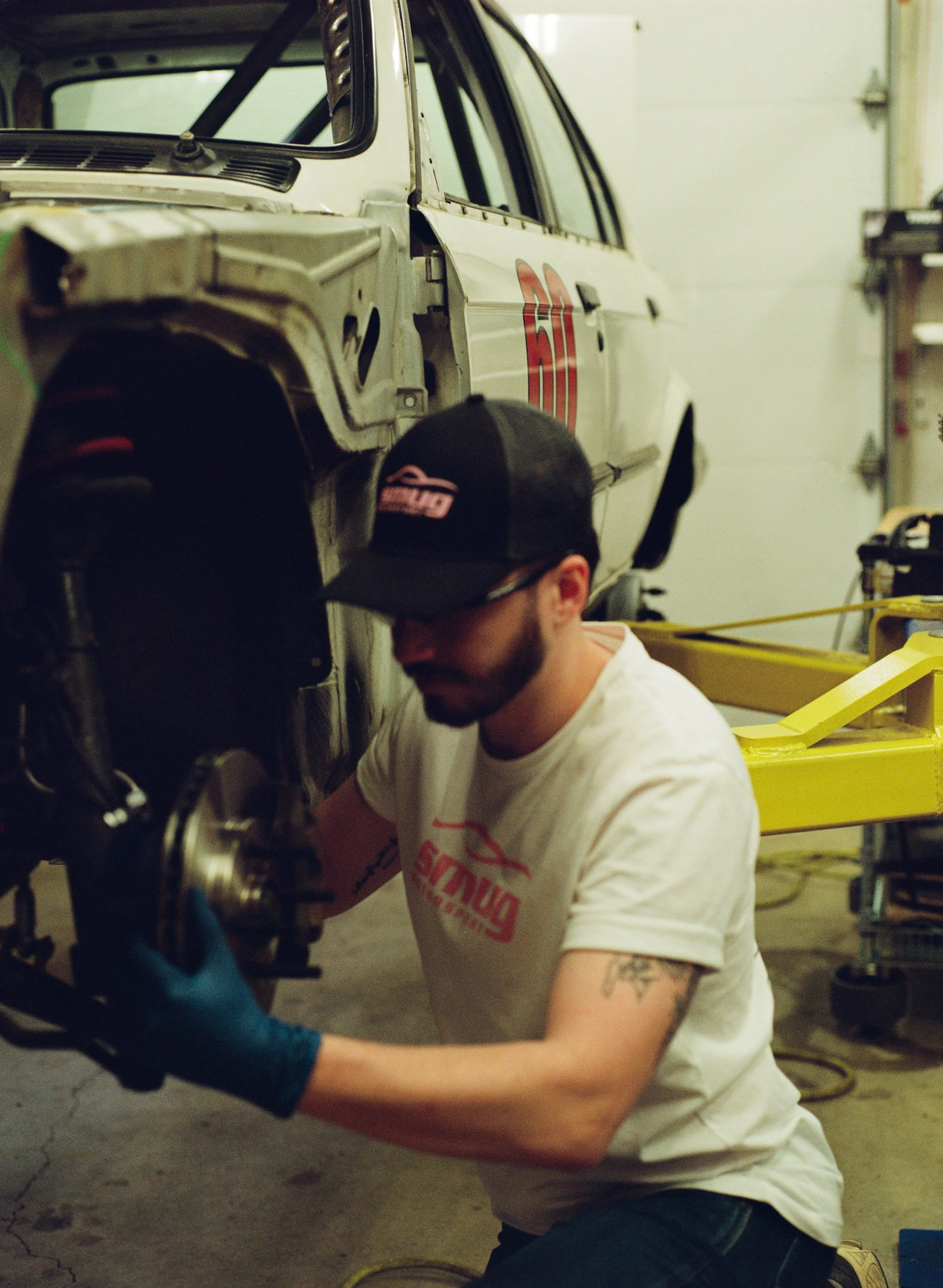 A person working on a vintage race car in a garage, wearing a black cap and white t-shirt. The race car has the number 20 on its side and is missing some body panels, revealing internal parts. The garage has tools and equipment in the background.