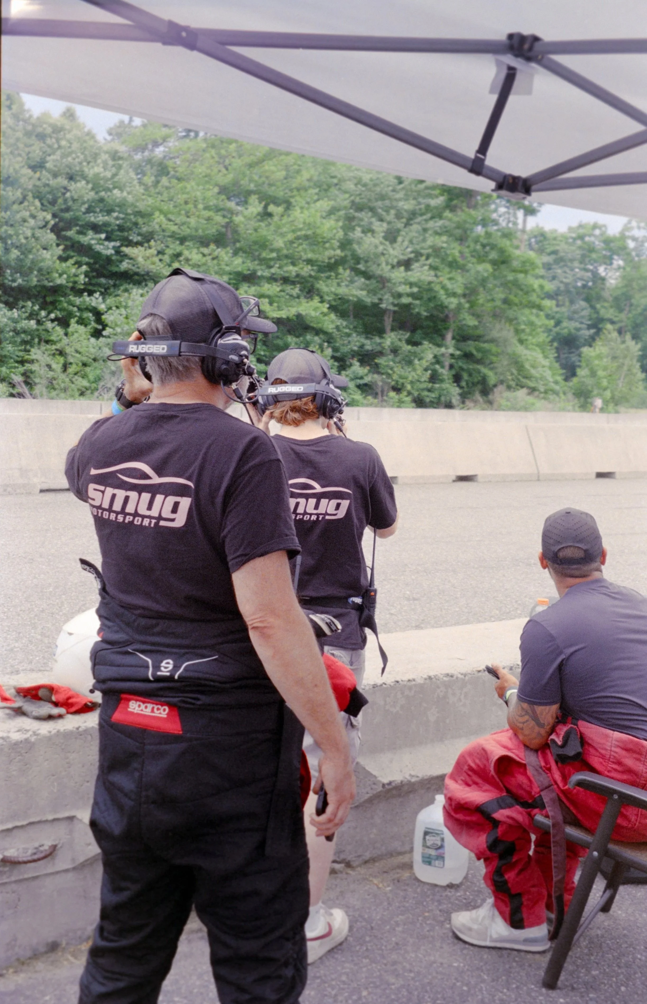 Group of people, likely race car drivers or pit crew, resting at an outdoor racing event with trees in the background. They are wearing racing team shirts and listening to communication headsets, some sitting on the curb and others standing.