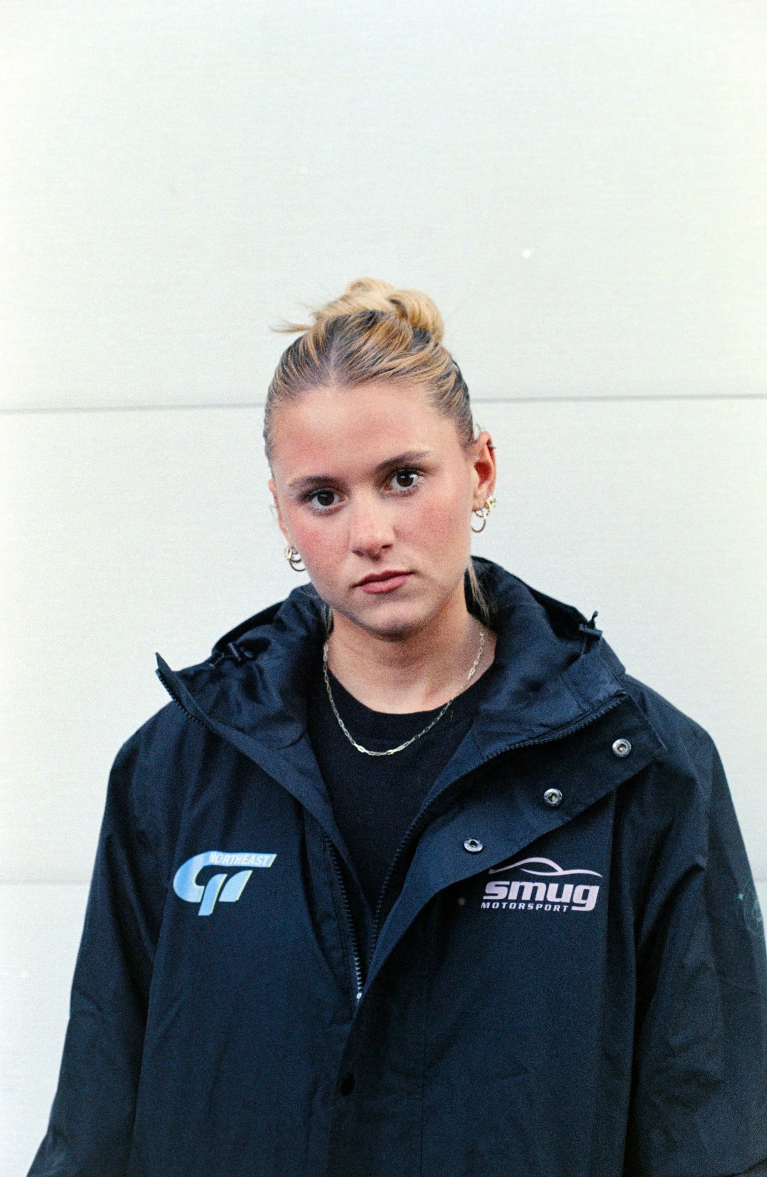 Young woman with blonde hair tied in a bun, wearing a black jacket with logos, standing against a plain off-white wall.