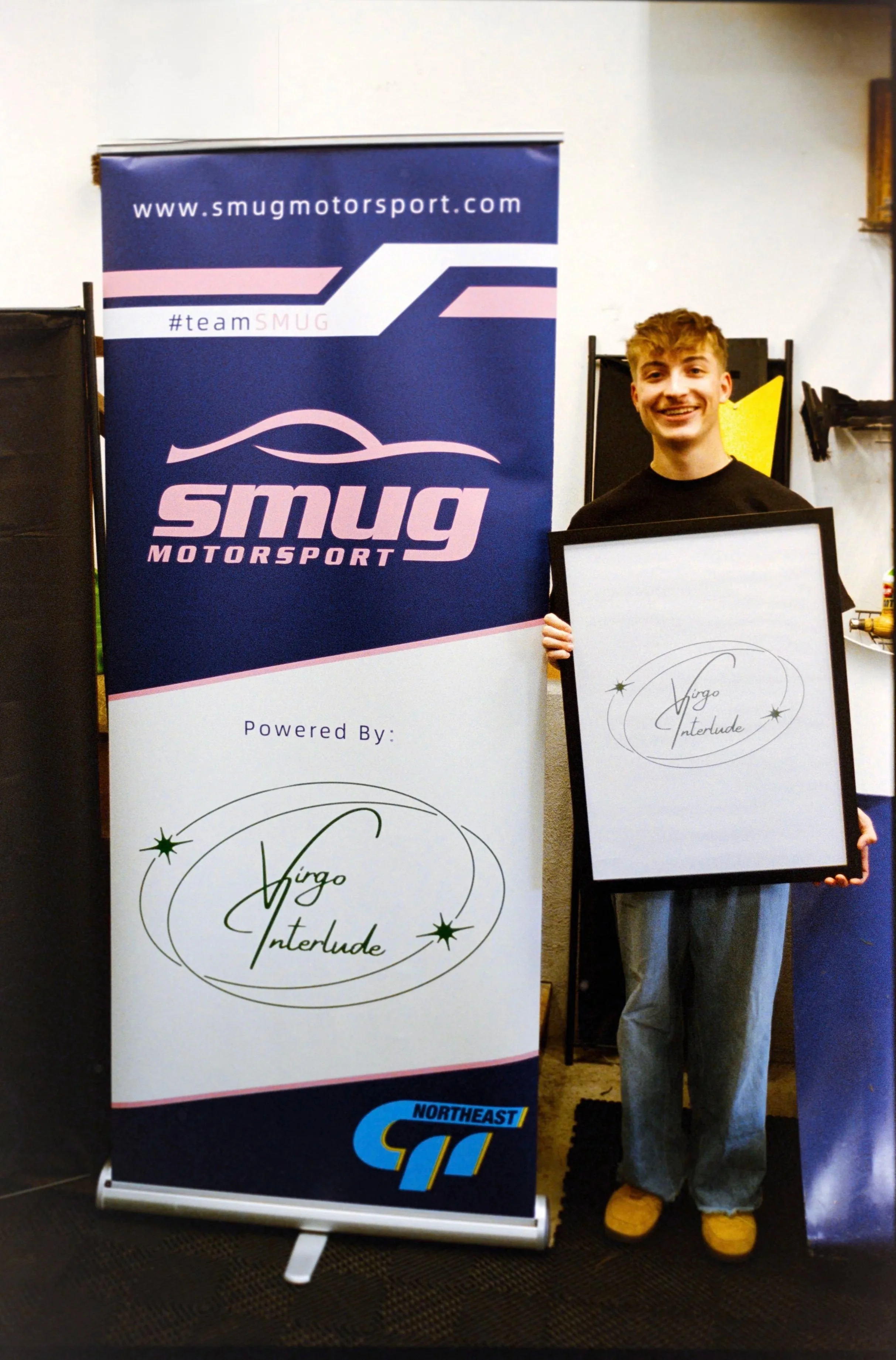 A person standing next to a Smug Motorsport banner and holding a framed sign that reads 'Virgo Interlude.' They are wearing gray pants, yellow shoes, and a black shirt.