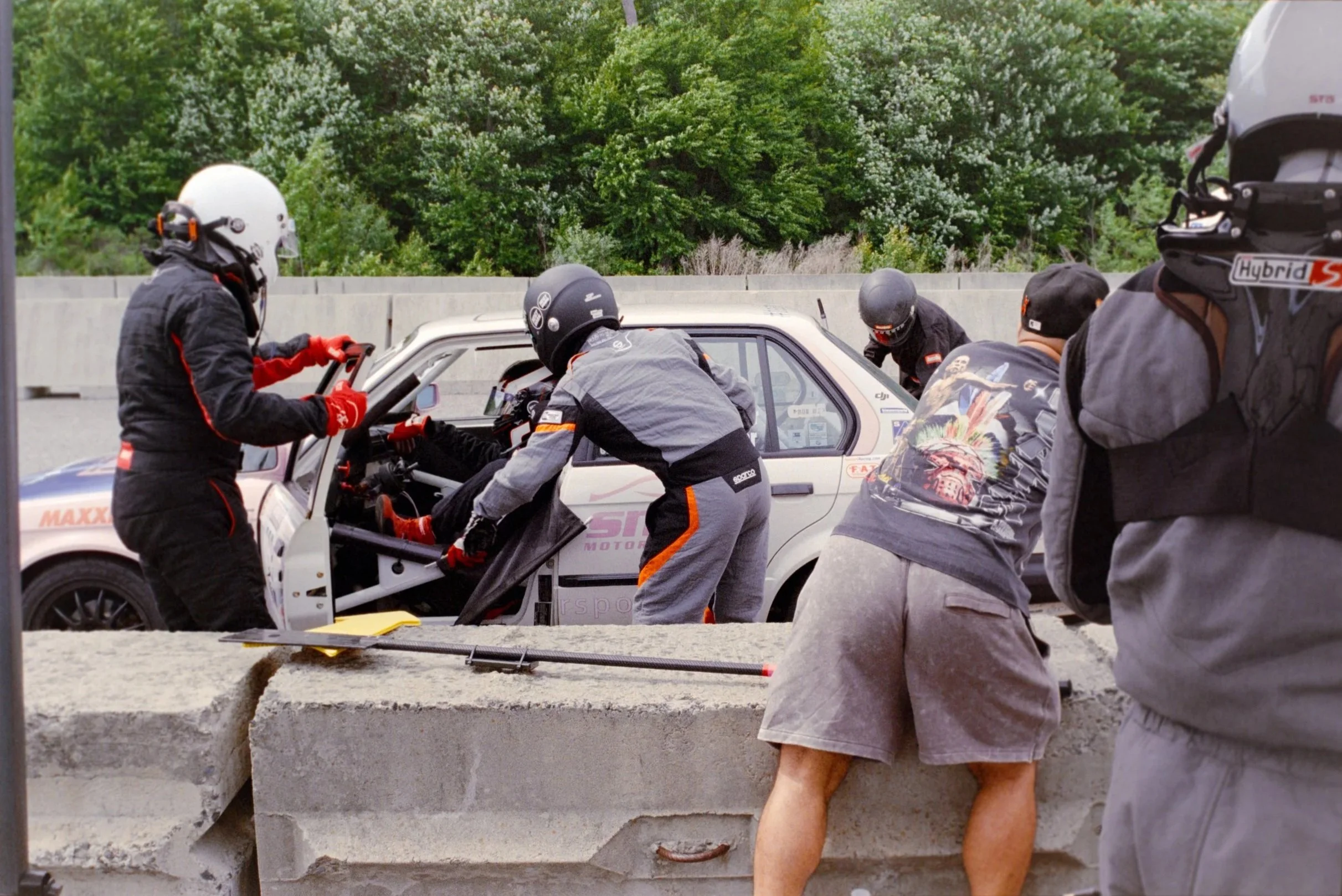 Race car team members working on a white race car during a pit stop, with greenery in the background.