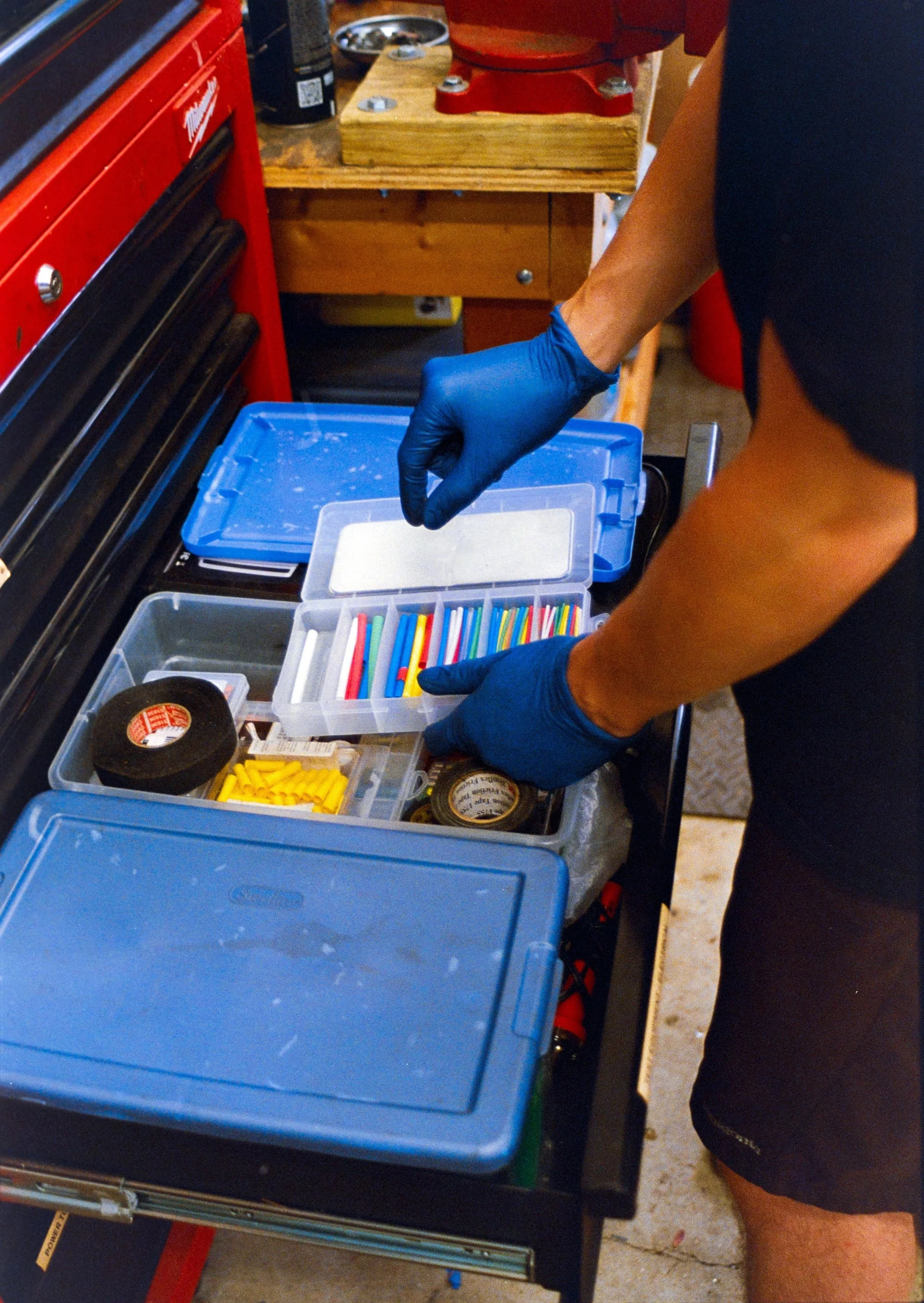 A person wearing gloves organizing plastic containers filled with colorful cable or wire connectors on a workbench in a workshop. The workbench has various tools and supplies, including black tape, a roll of yellow cable ties, and other miscellaneous