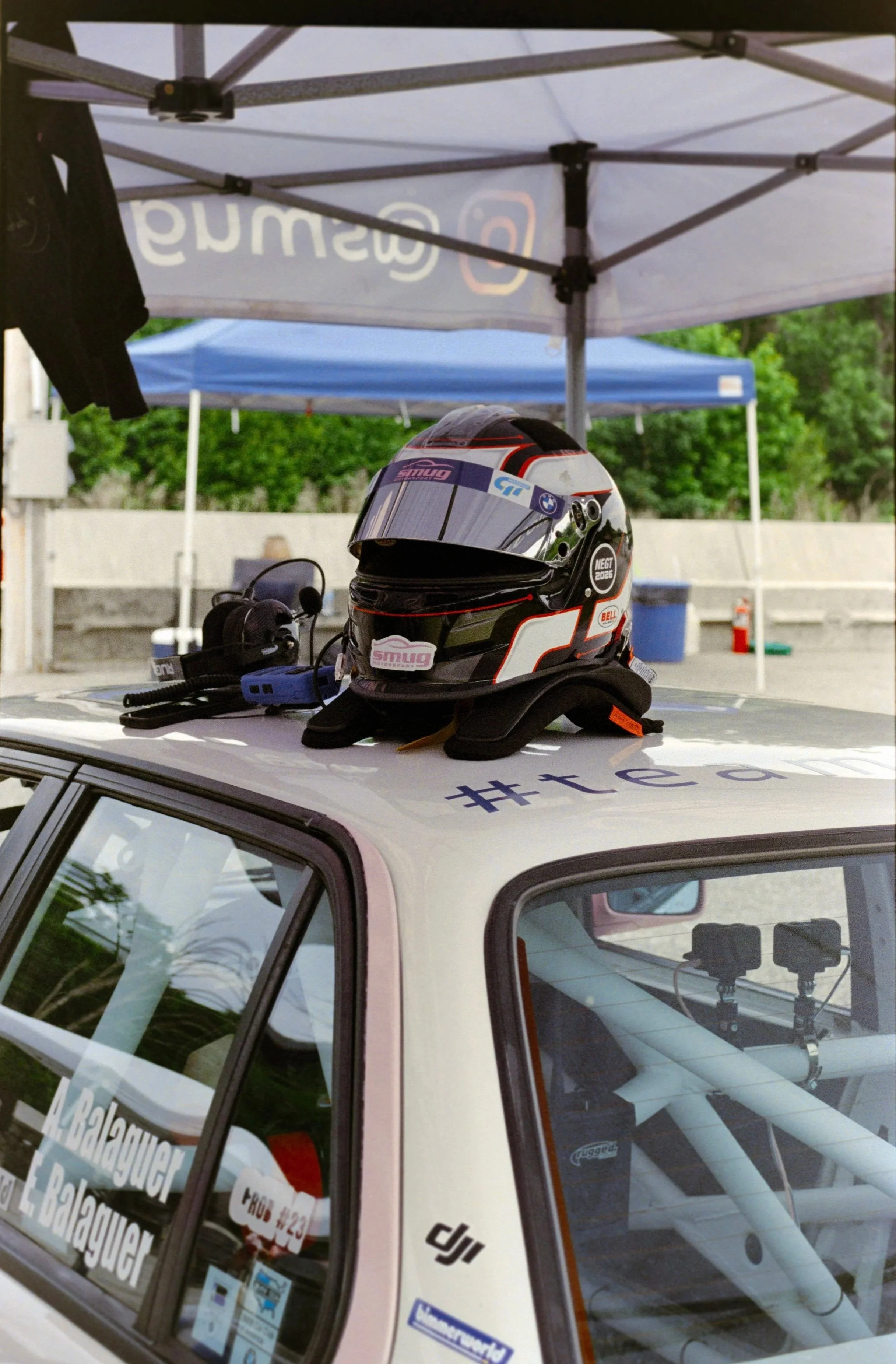A racing helmet with a black, red, and white design resting on top of a white vehicle. The helmet is adjacent to a blue and gray canopy tent, with trees and another canopy visible in the background.