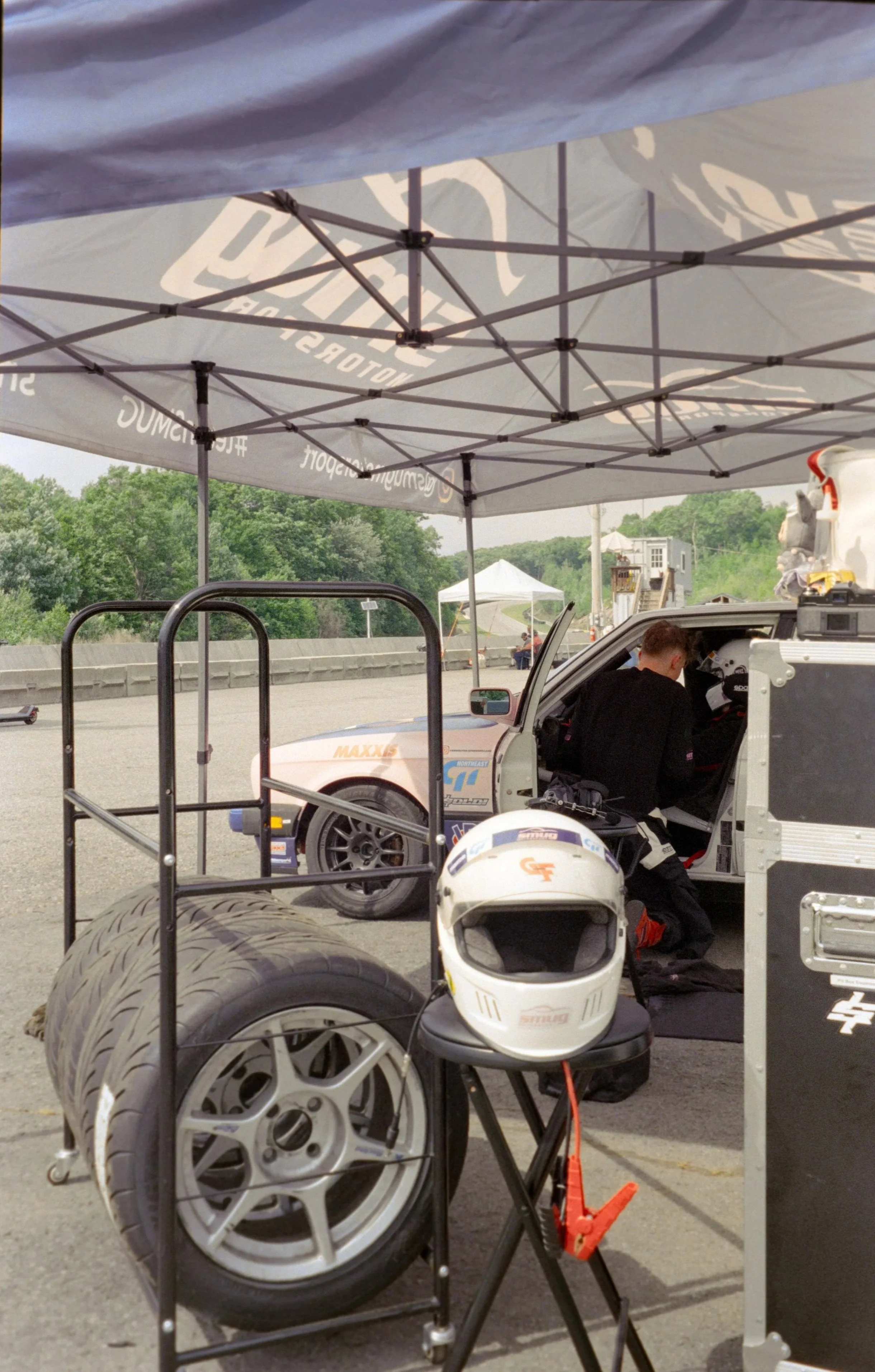 A racing pit area with a canopy tent, racing tires, a race helmet, and a person working on a race car.