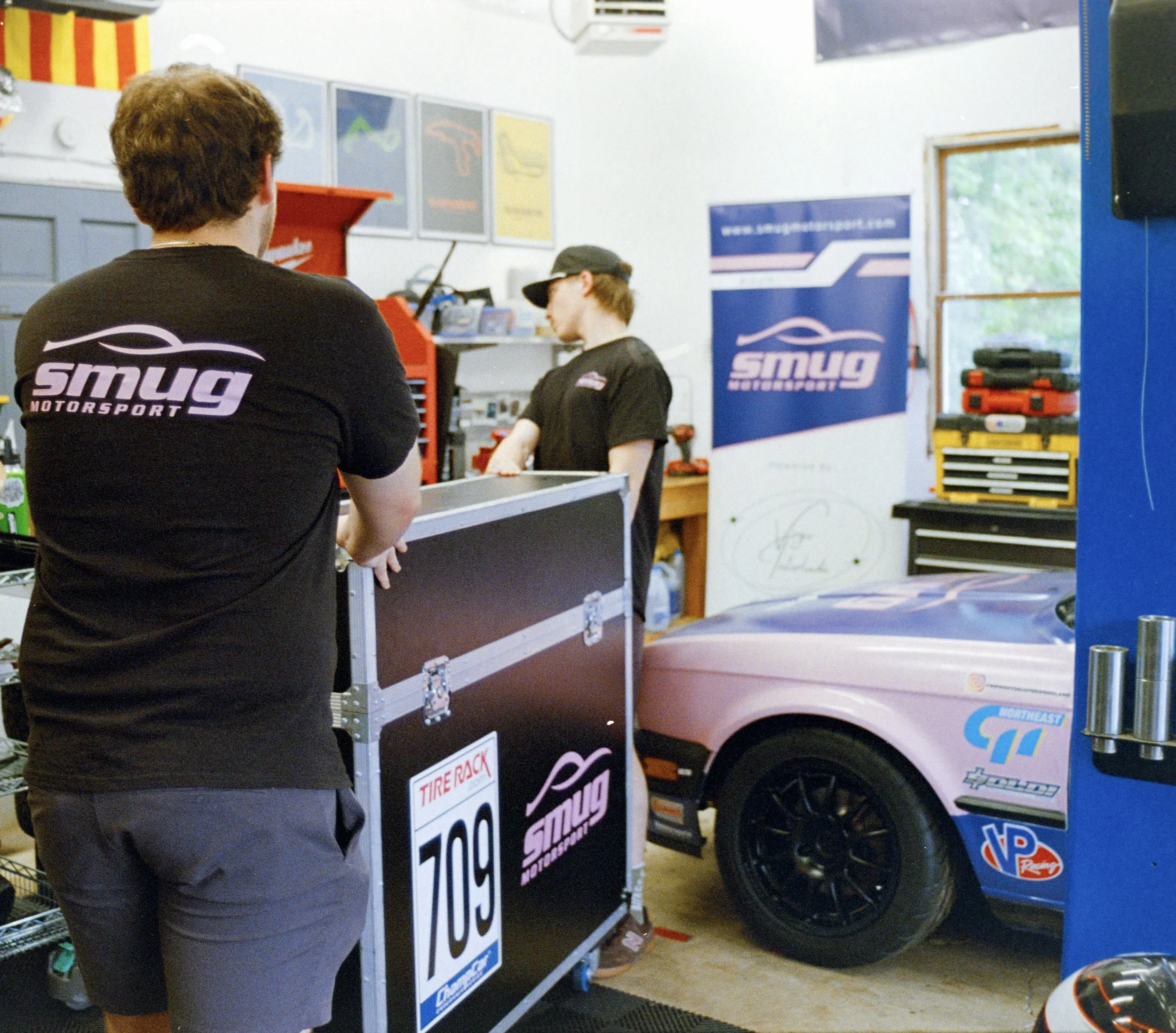 People working at a motor sports workshop with signage displaying prices and logos, including a 'Smug Motorsports' banner and a vehicle with sponsor decals.