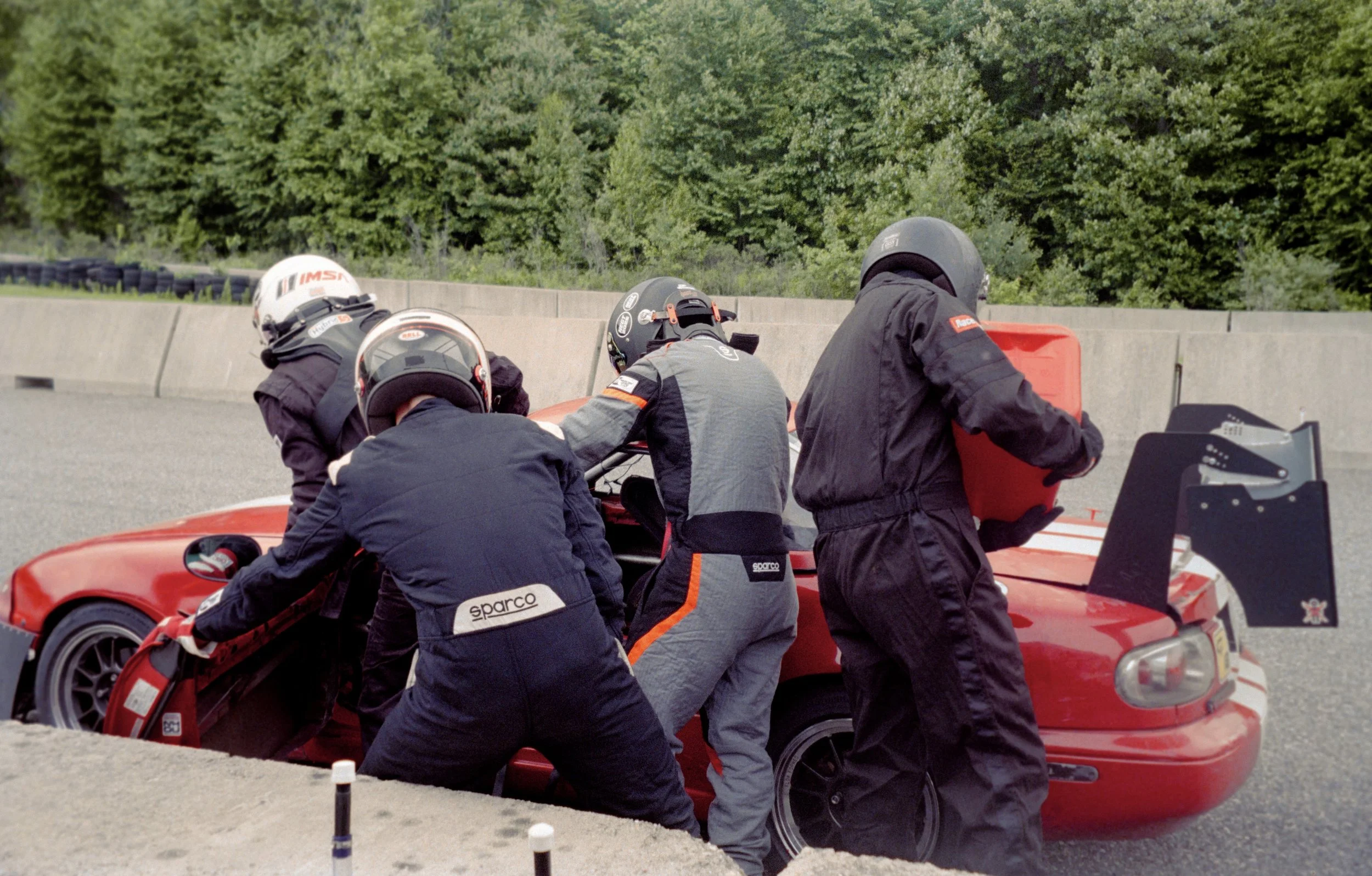 A team of race car mechanics working on a red sports car at a race track, with green trees in the background.