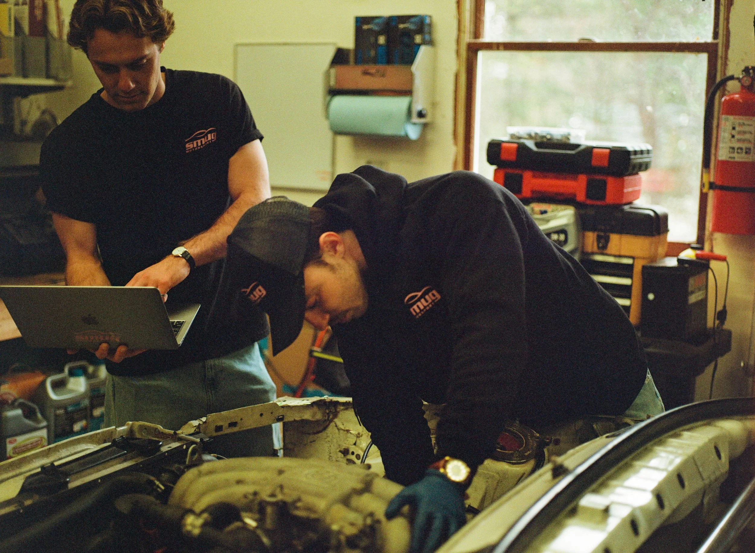 Two men working on a car engine in a garage, with tools and equipment around them.