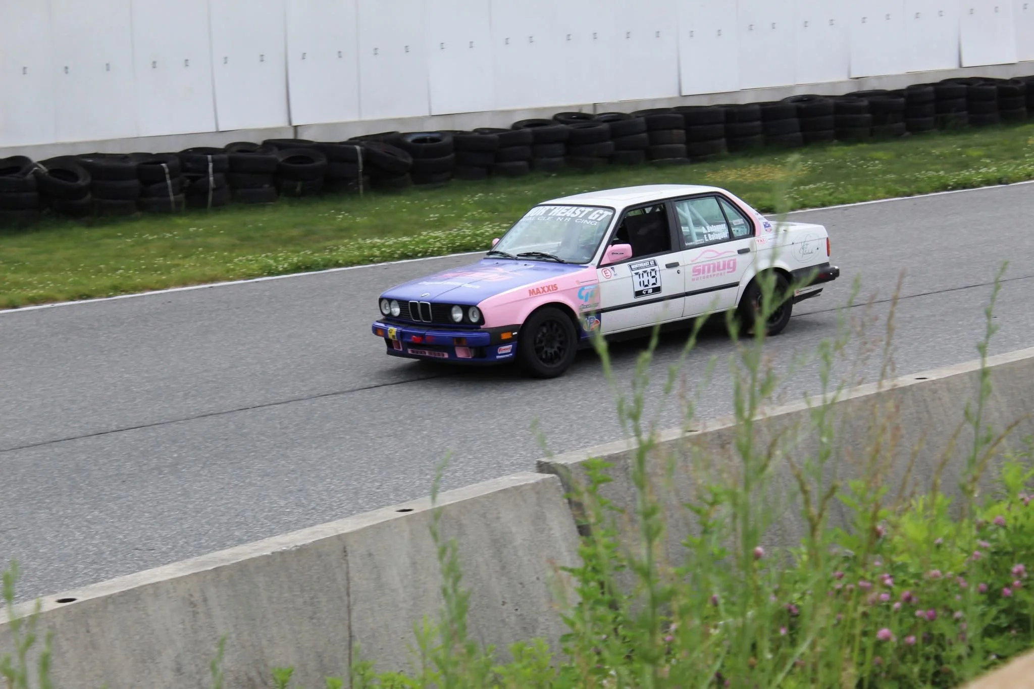 A vintage race car on a race track, surrounded by a barrier of black tires and a white wall with supervision markings.