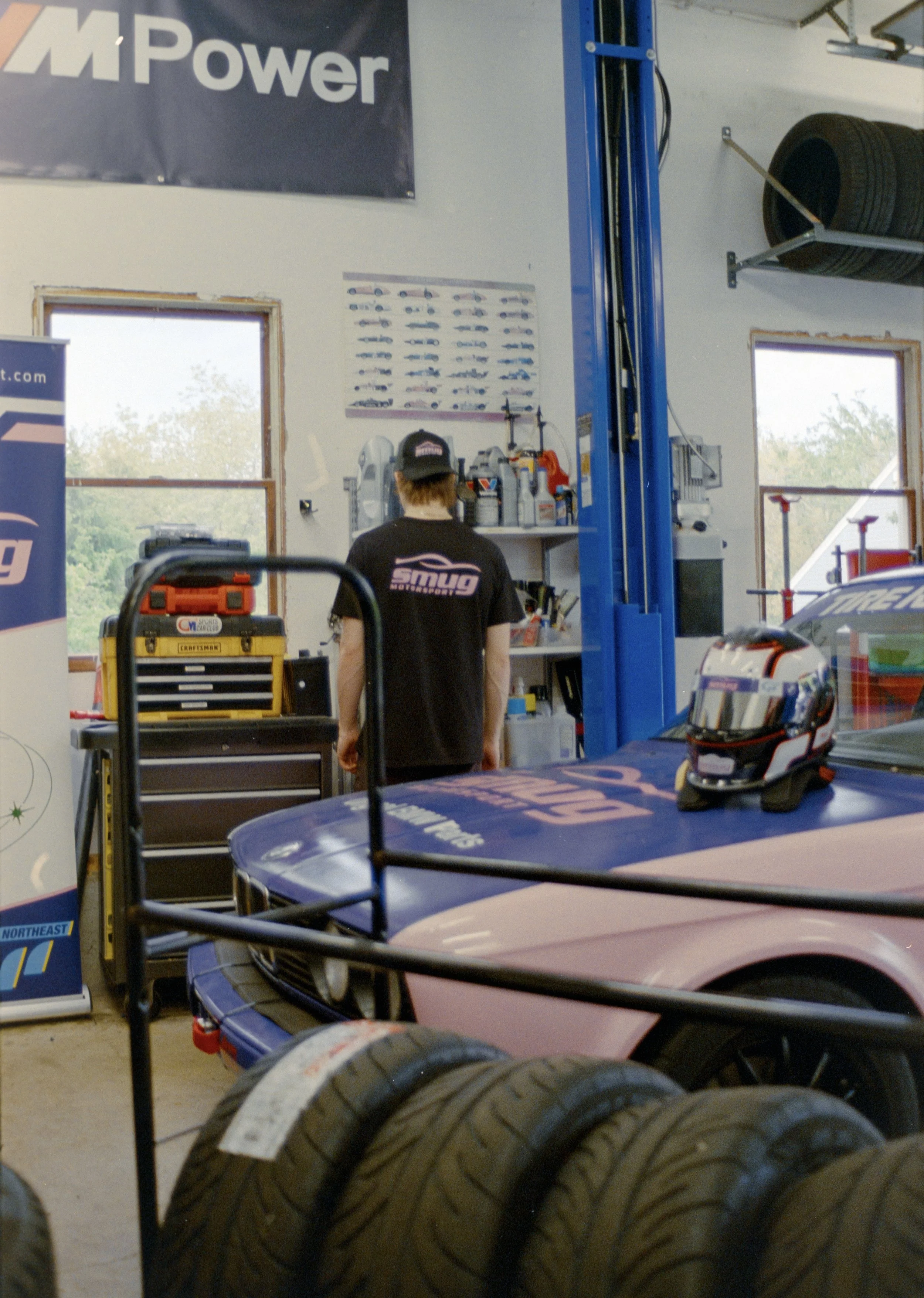 Inside an automotive workshop with tires stacked on racks, a person working at a station, and racing cars with helmets on a table.