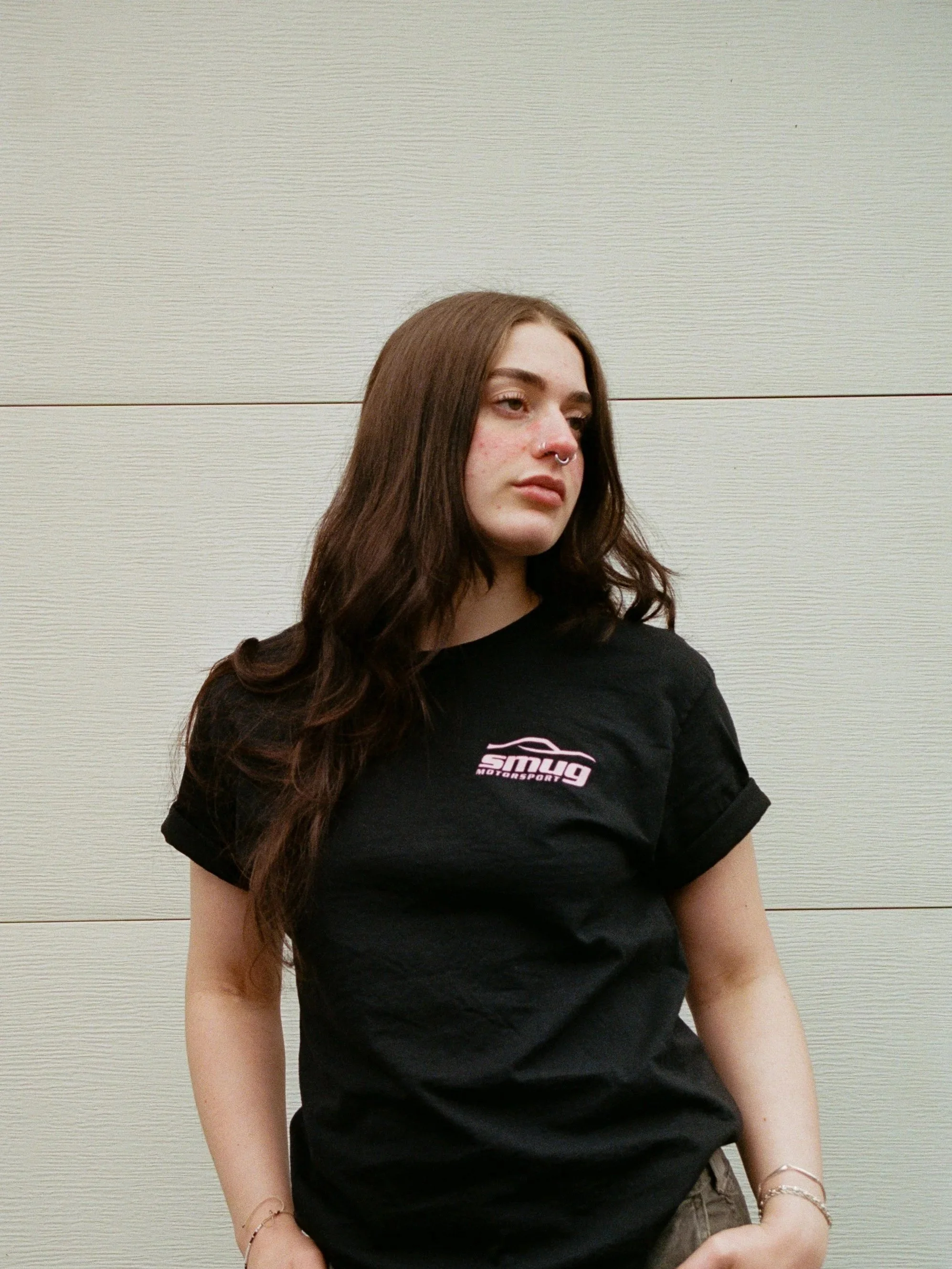 A young woman with long brown hair and a septum piercing standing against a plain wall, wearing a black t-shirt with a pink and white logo.