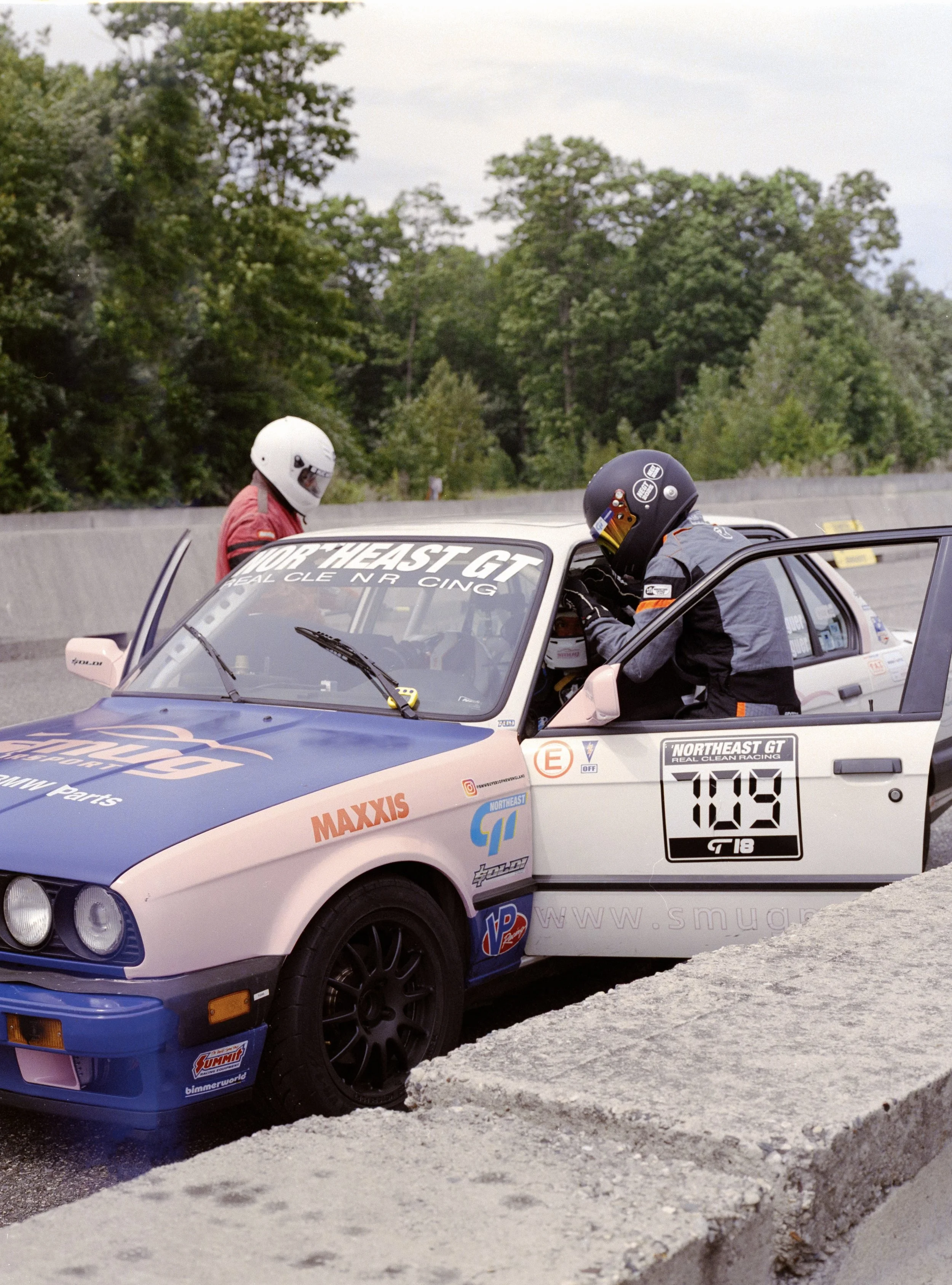 Race car with two mechanics wearing helmets working on the vehicle on a race track surrounded by trees in the background.
