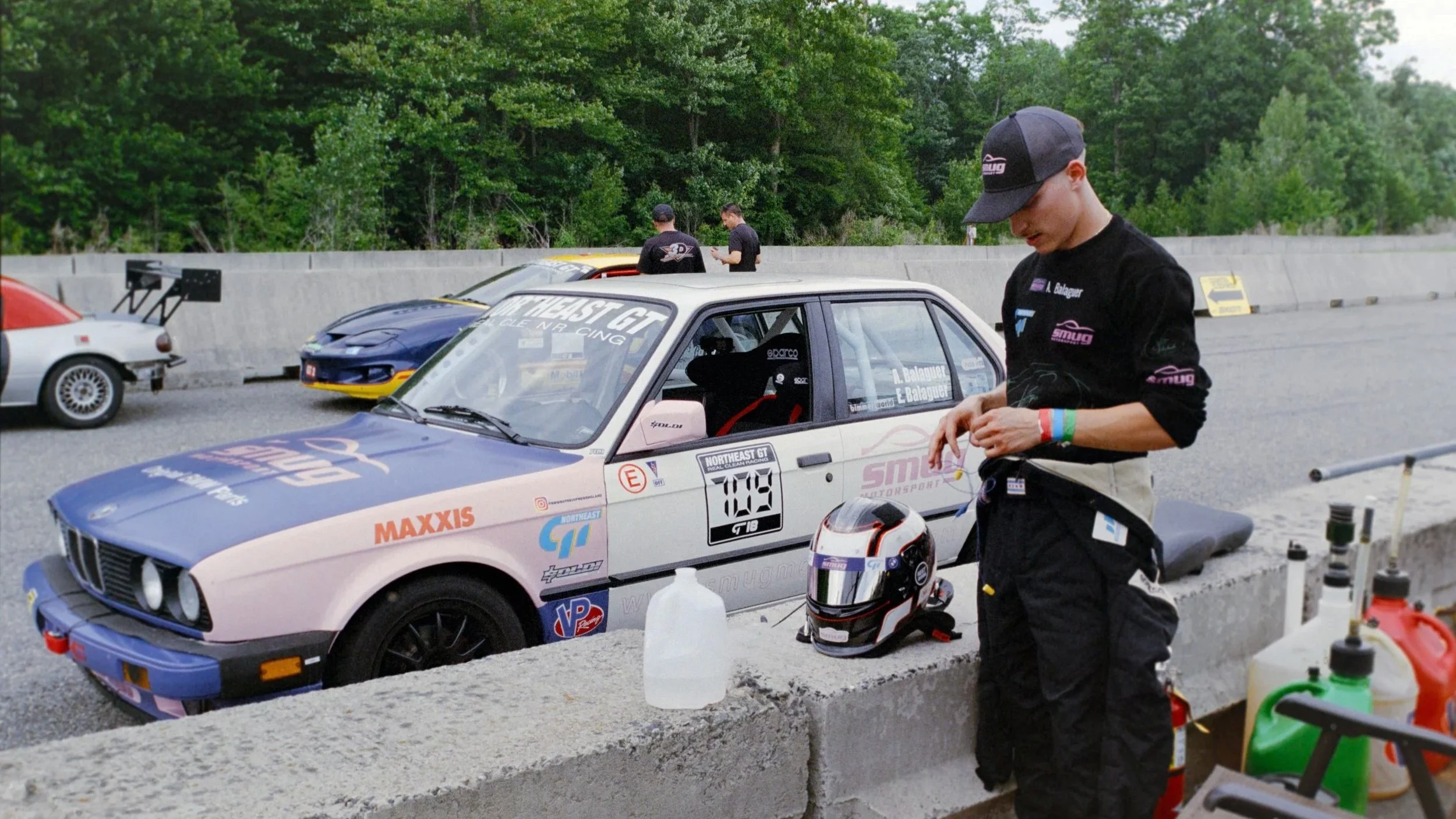 Race car driver in racing suit standing next to a race car in a pit stop area, with other race cars and crew members in the background.