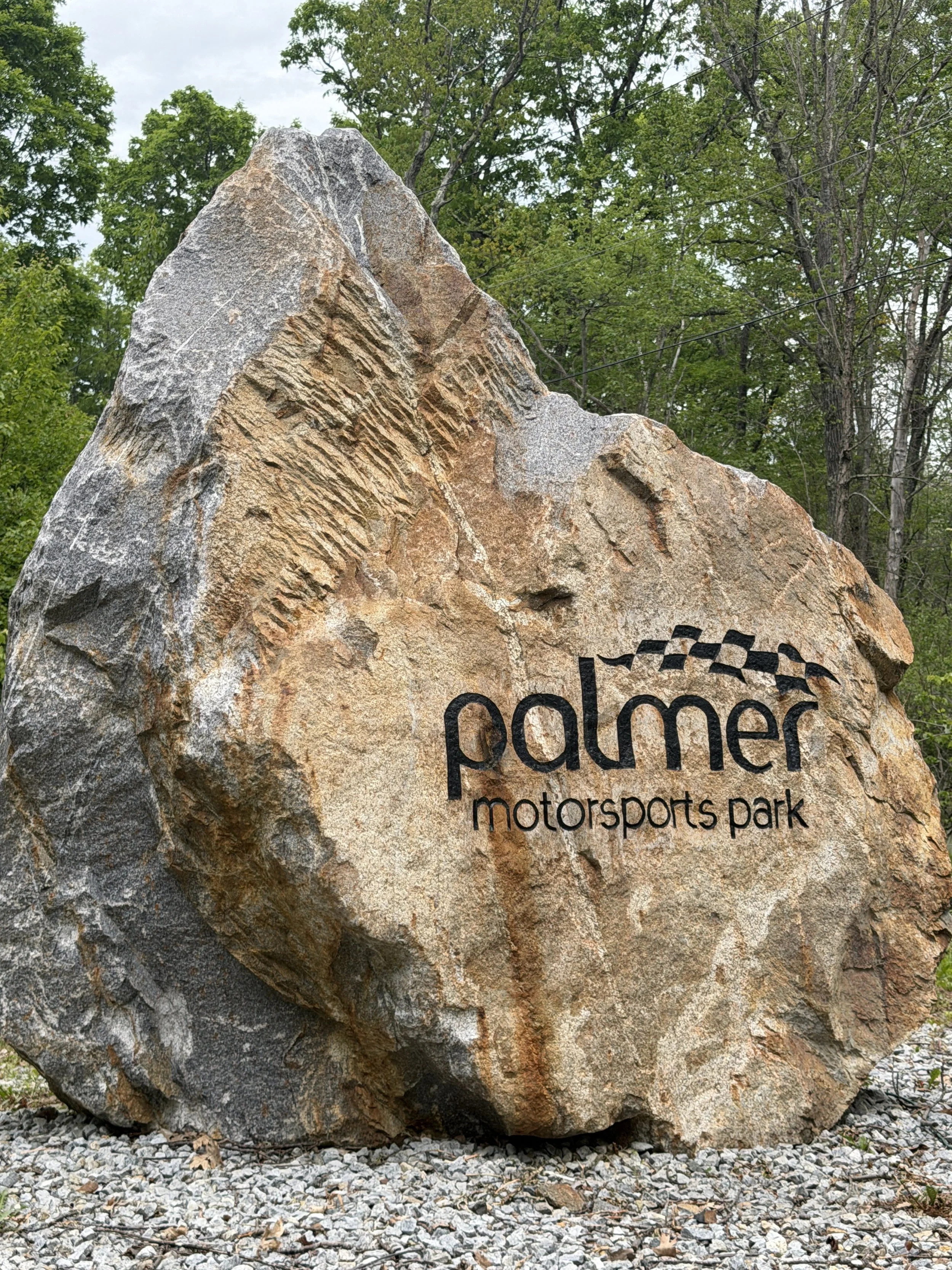 Large rock with 'Palmer Motorsports Park' logo engraved on it, surrounded by small rocks and trees in the background.