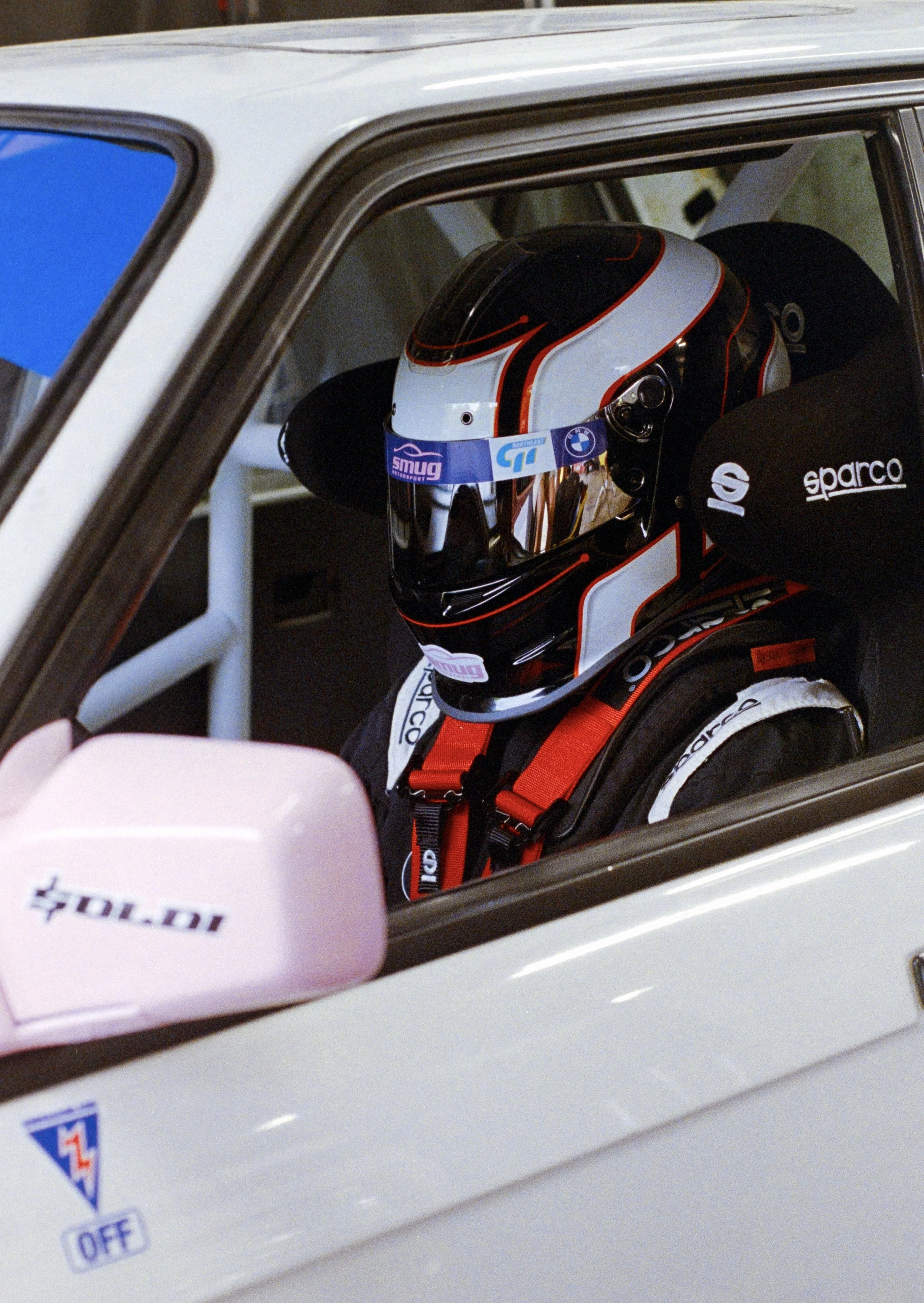 A person sitting inside a white race car wearing a black helmet with red and white accents and a racing suit with red straps, visible through the car window.