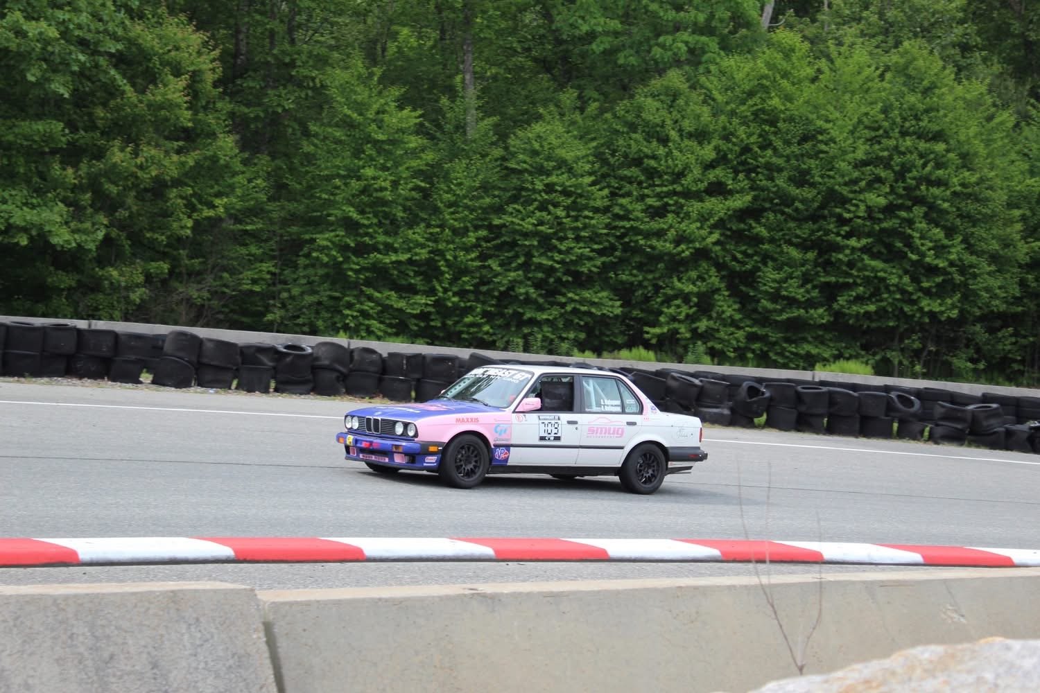 A pink and white vintage BMW racing car on a race track surrounded by green trees and black tire barriers.