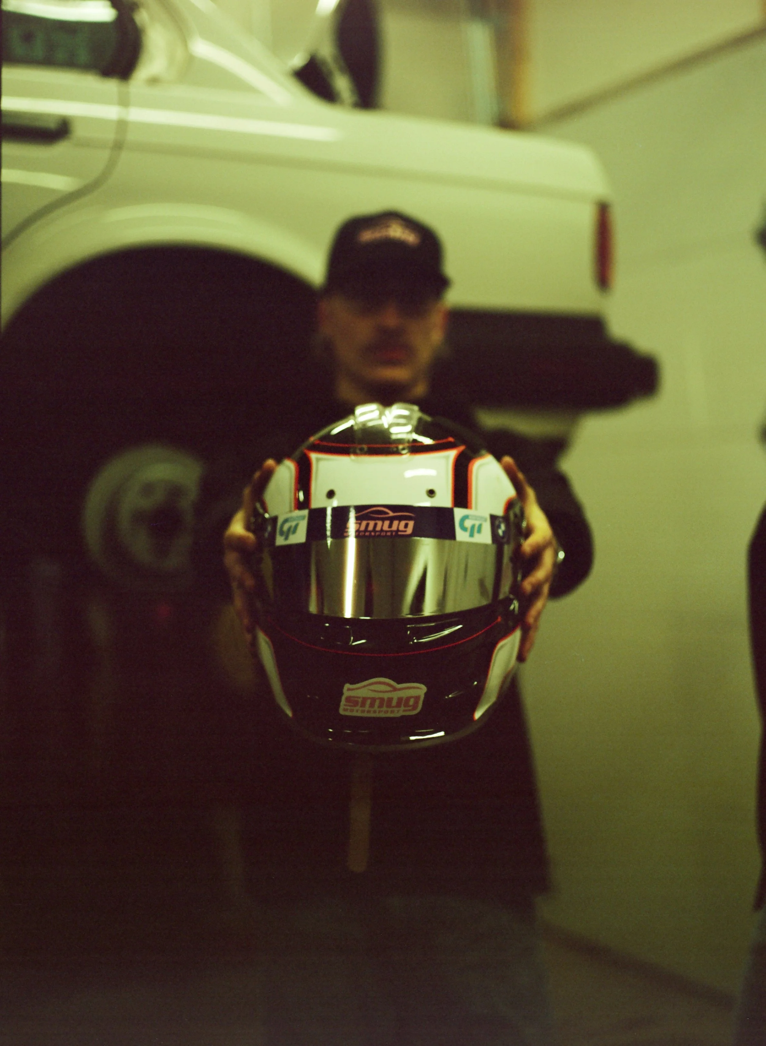 A person taking a mirror selfie holding a racing helmet with various sponsor logos, wearing a dark shirt and a cap, standing in a dimly lit room with a vintage car reflection in the mirror.