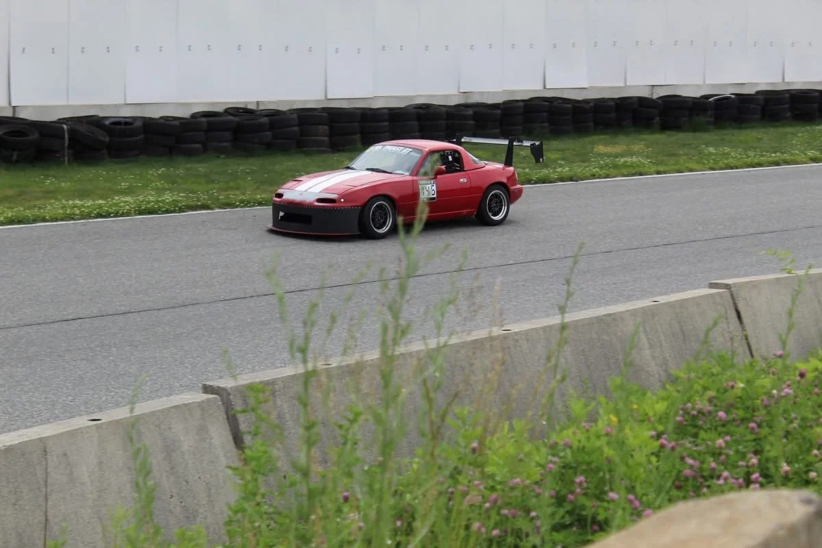 A red race car with a black rear wing on a race track, with tire barriers and a white wall in the background.