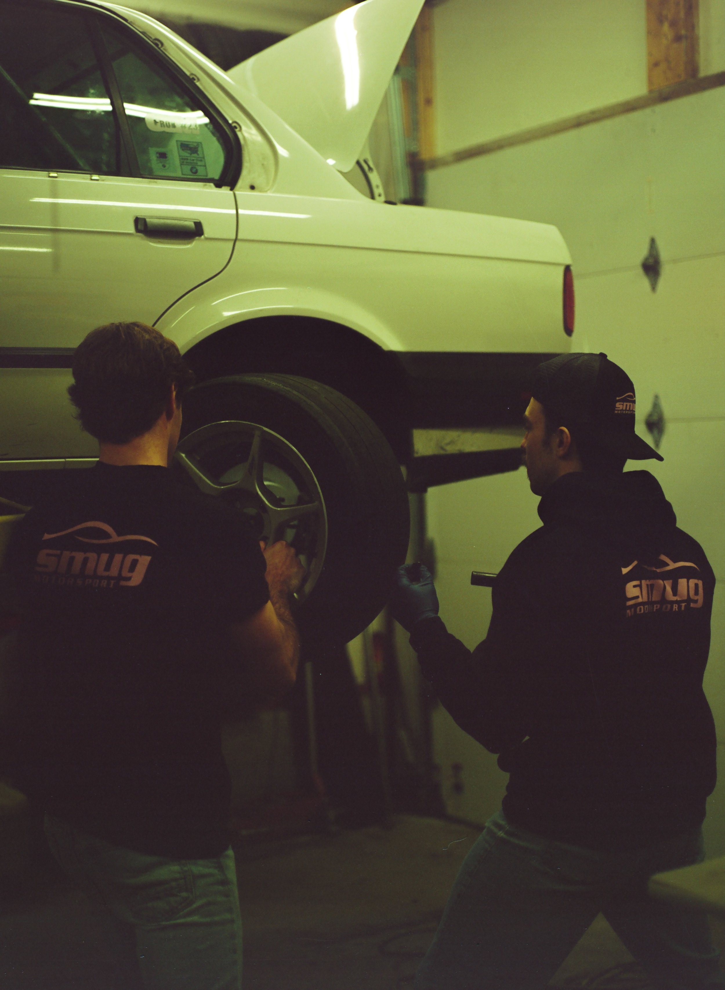 Two men in black shirts and one boy in a black hoodie working on a white classic car inside a garage or workshop.