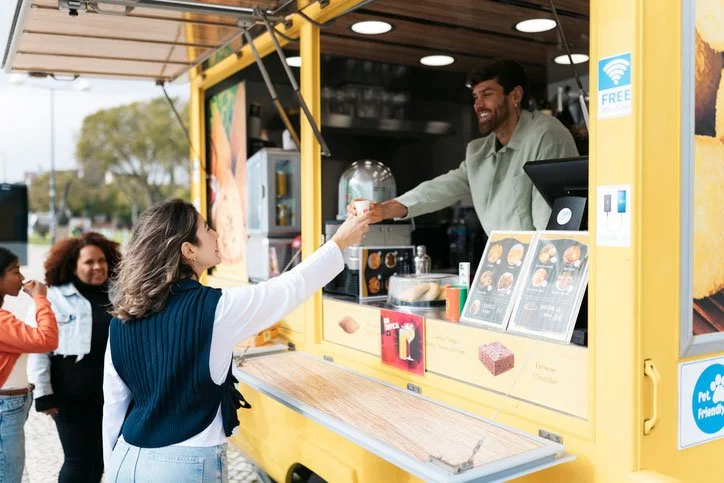 A woman in a navy vest and white shirt is receiving a drink from a smiling man in a food truck. The food truck is yellow with menus displayed, and people are waiting in line outside.
