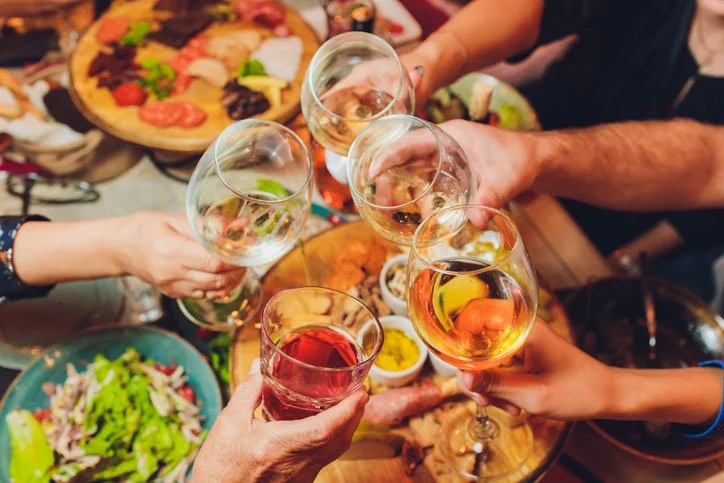 People clinking wine glasses with a pizza, seafood, salad, and dips on a table.