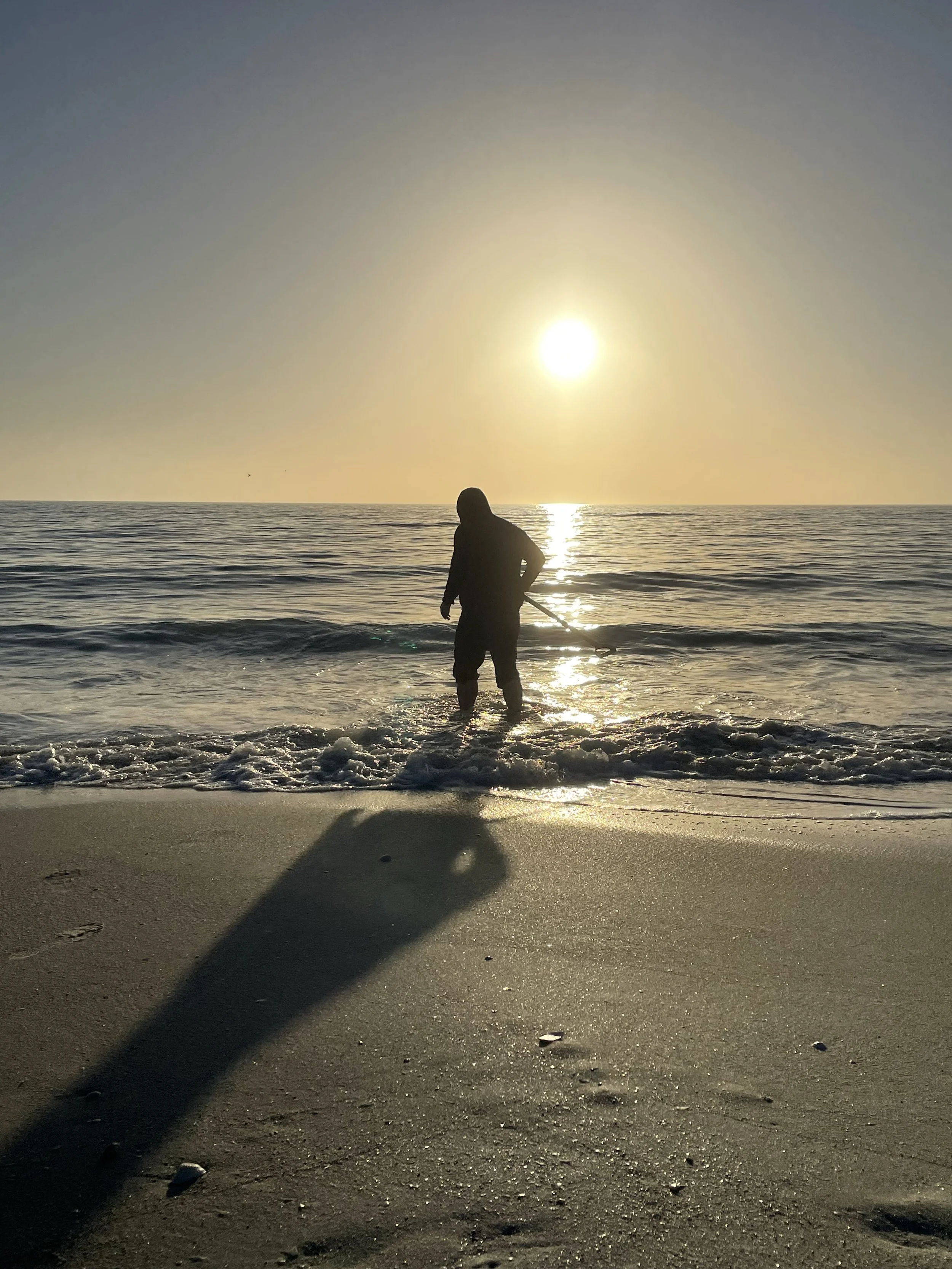 Person standing in shallow ocean water during sunset or sunrise at the beach, casting a long shadow on the sand.  Metal Detecting in the surf.