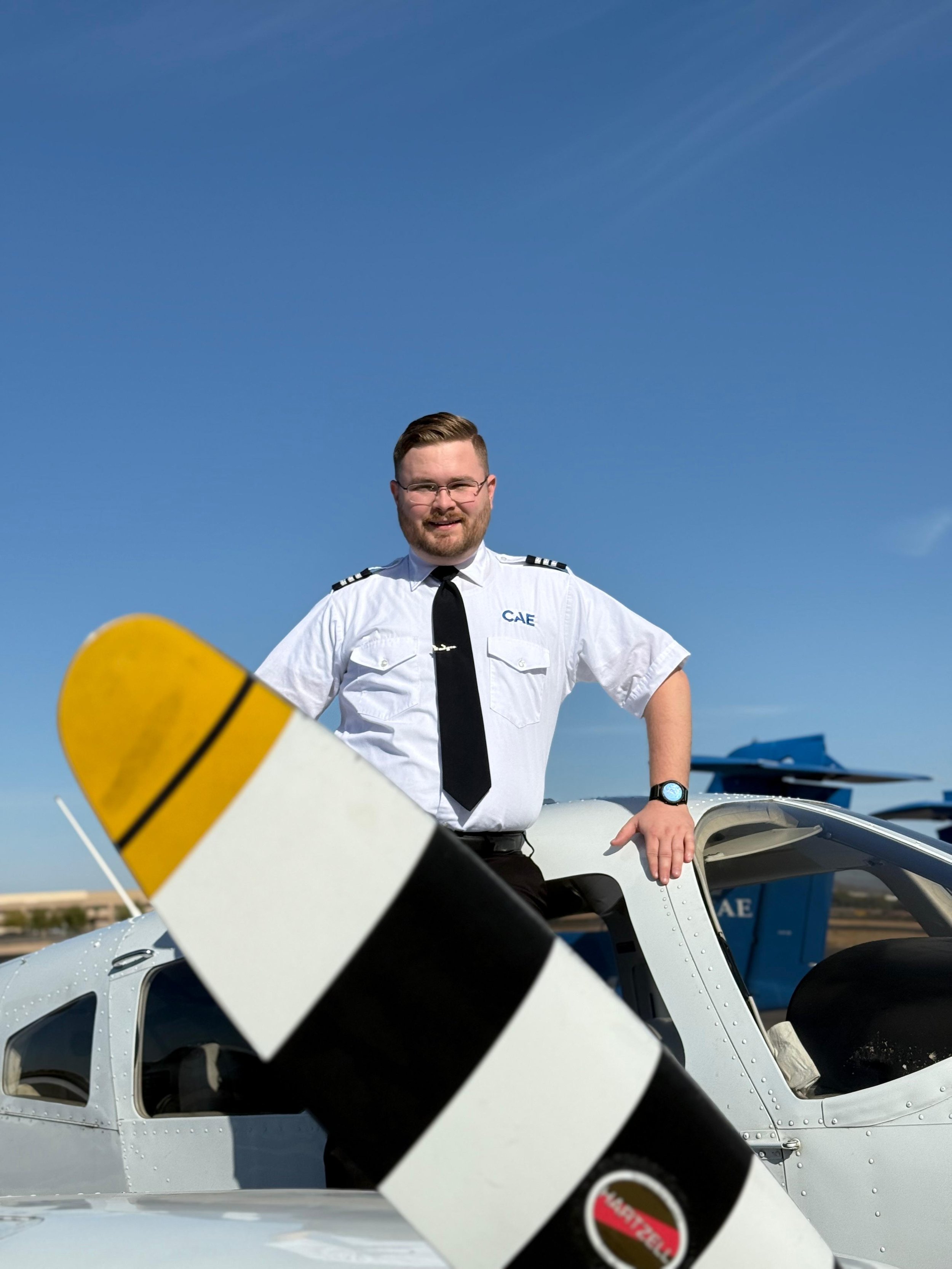 A smiling man in a white pilot uniform with black epaulets and a black tie standing next to a small white airplane. The man is leaning on the airplane's open cockpit door, and there are airport tarmac and other planes in the background. The sky is clear and blue.