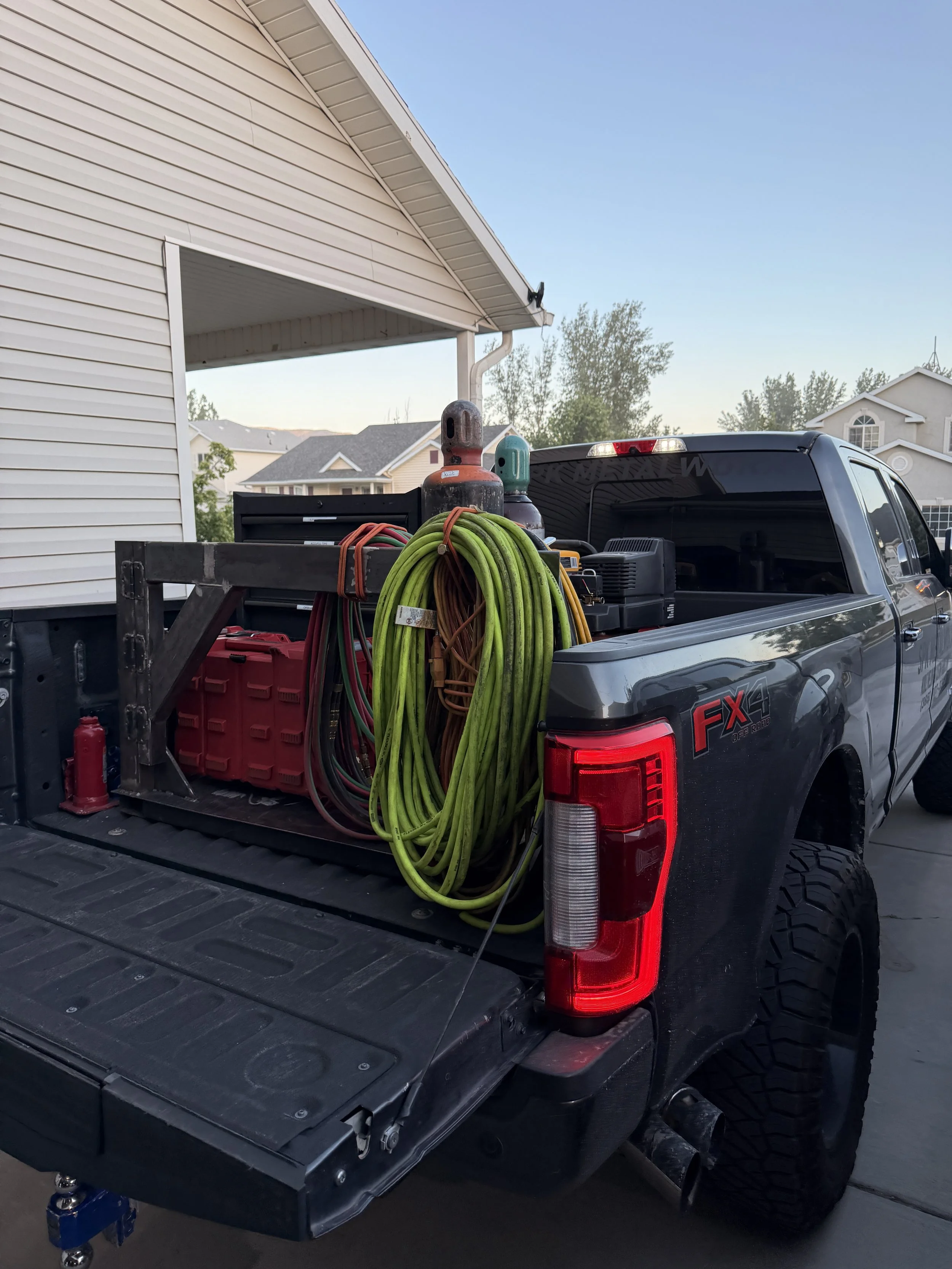 The image shows the back of a gray pickup truck loaded with various tools and equipment, including coiled extension cords, a propane tank, and a power generator, parked in a residential driveway next to a beige house with white siding, under a clear 