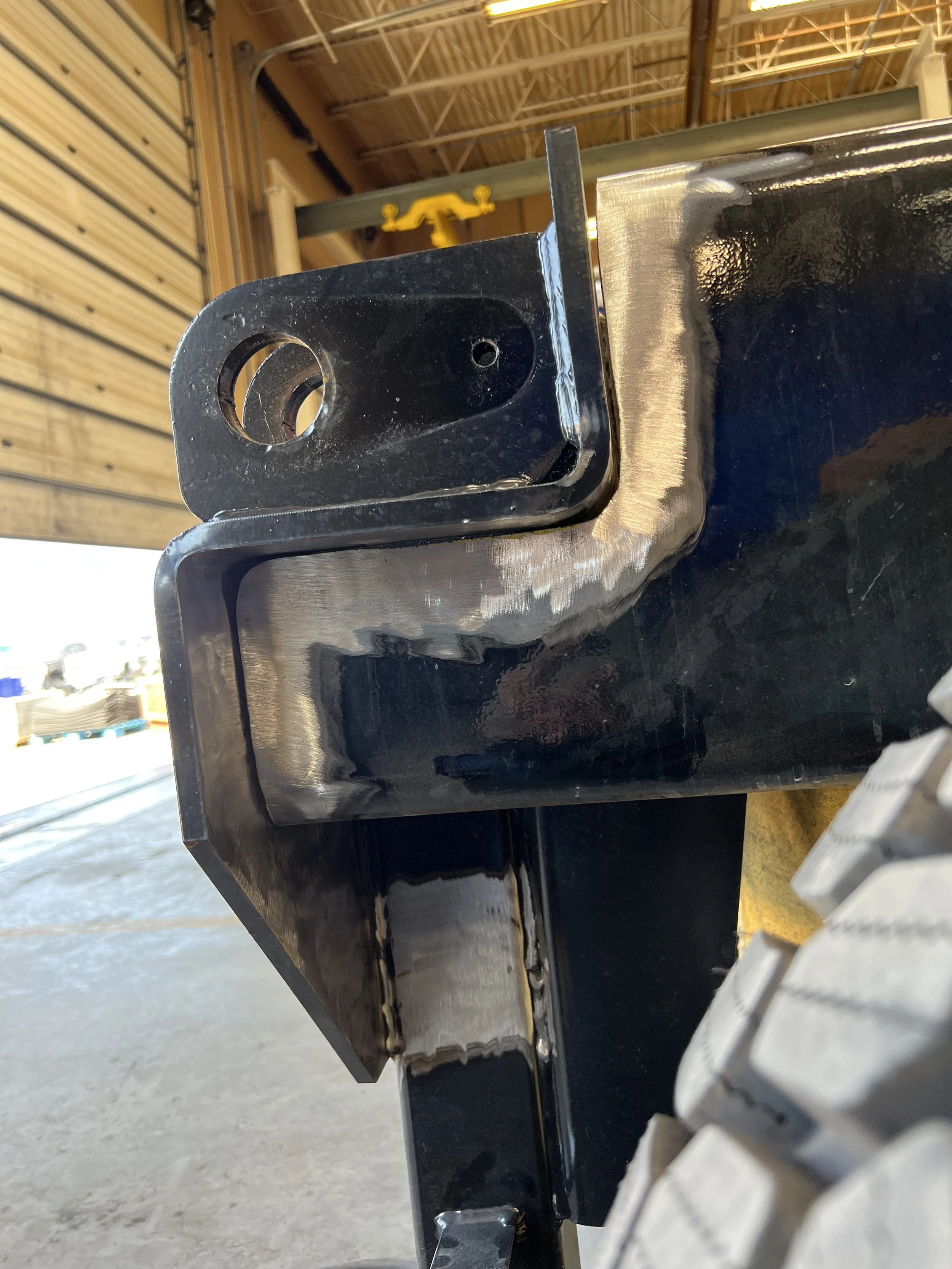 Close-up view of a black metal trailer hitch attached to the front of a vehicle, with part of a tire visible on the right side of the image, in an indoor setting with a high ceiling and some industrial equipment in the background.